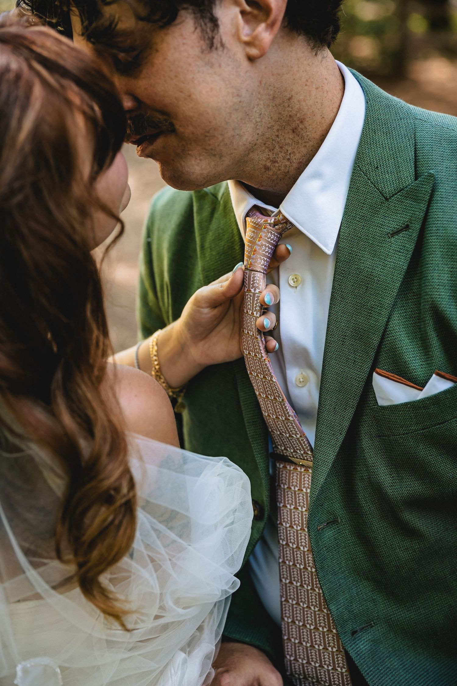 Bride in white tulle gown pulls groom's patterned tie as they share a romantic moment, groom wearing green suit.
