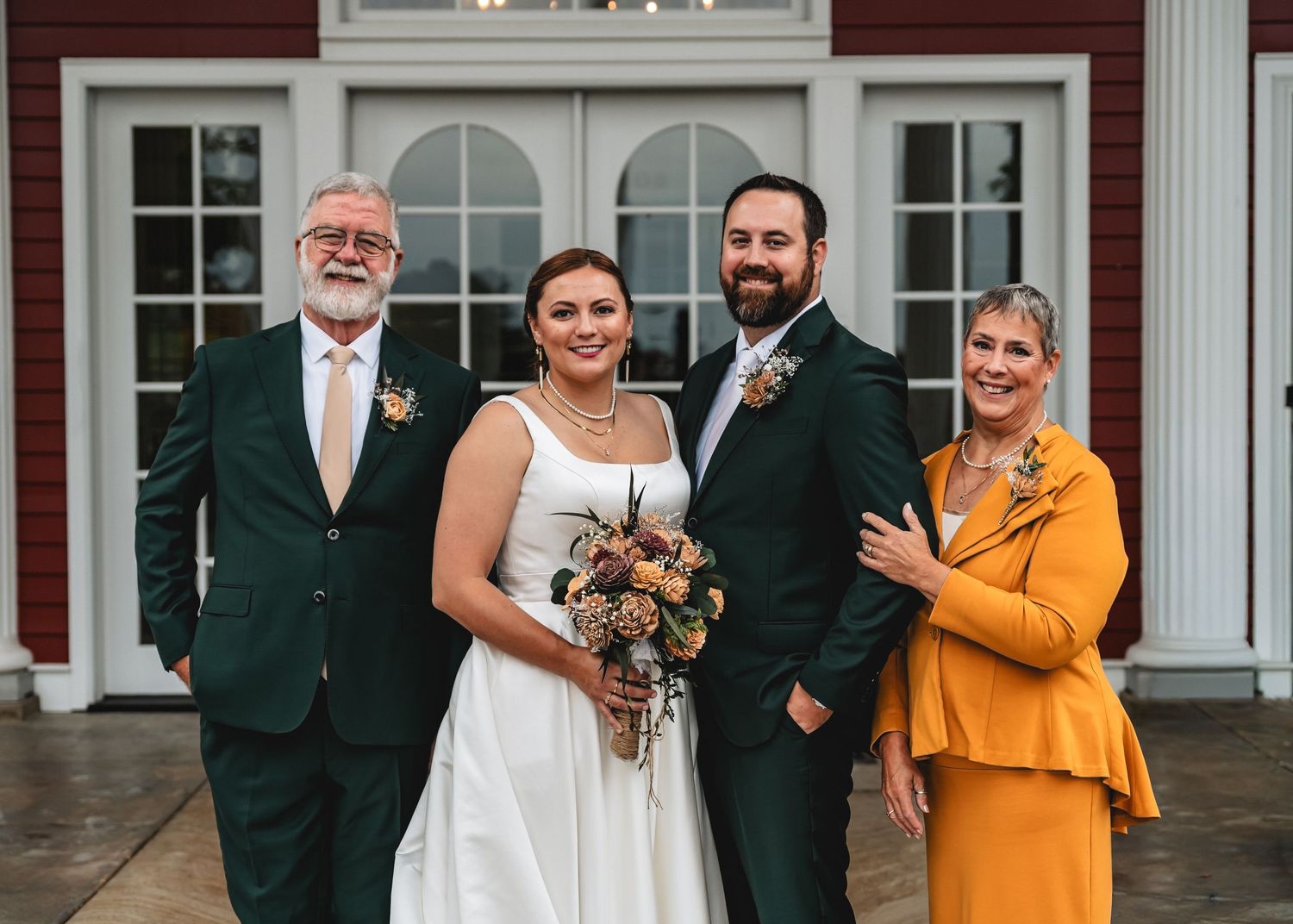 Wedding couple in white dress and green suit pose with family outside a white building with French doors.