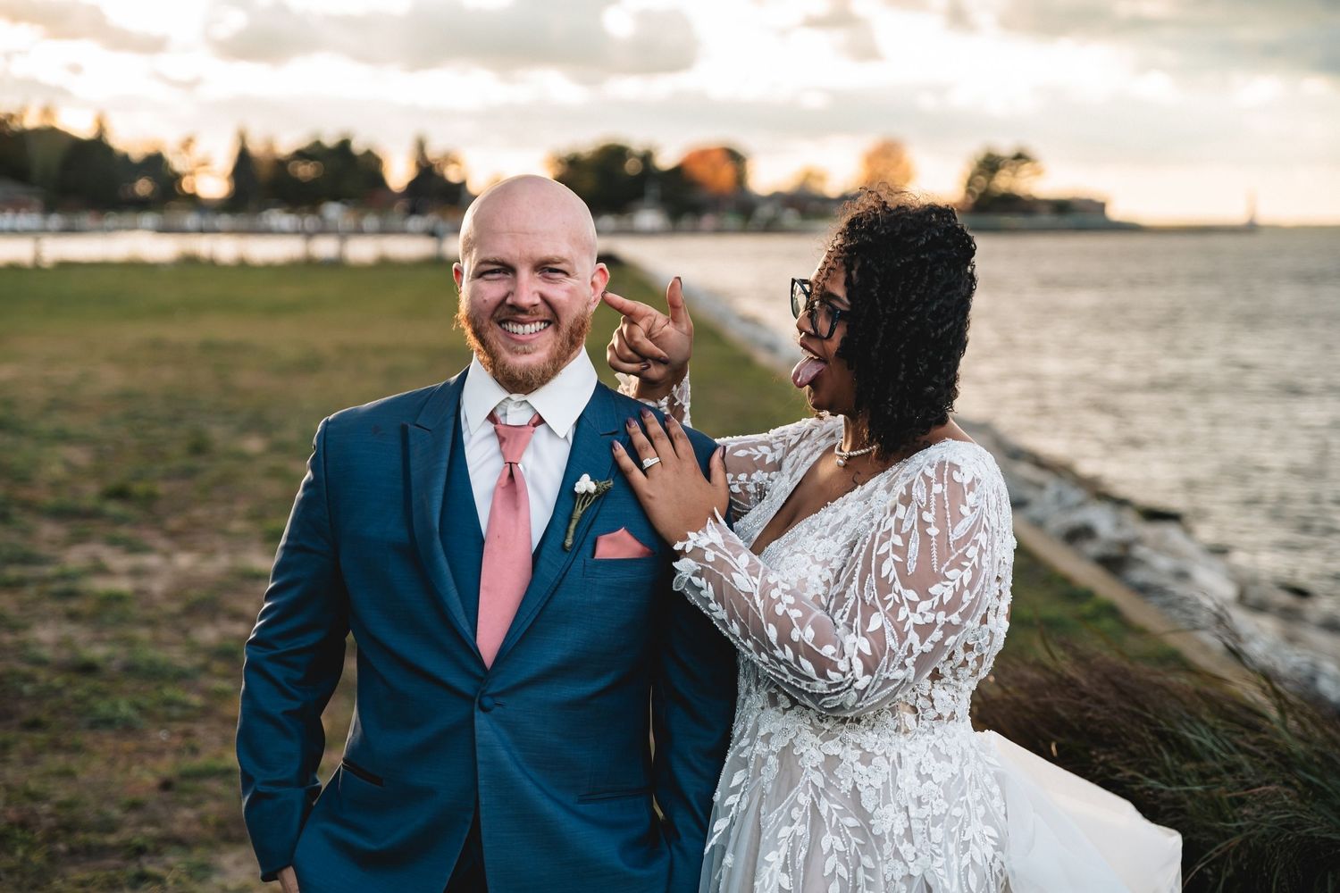 A couple in wedding attire shares a romantic moment at sunset by the water's edge.