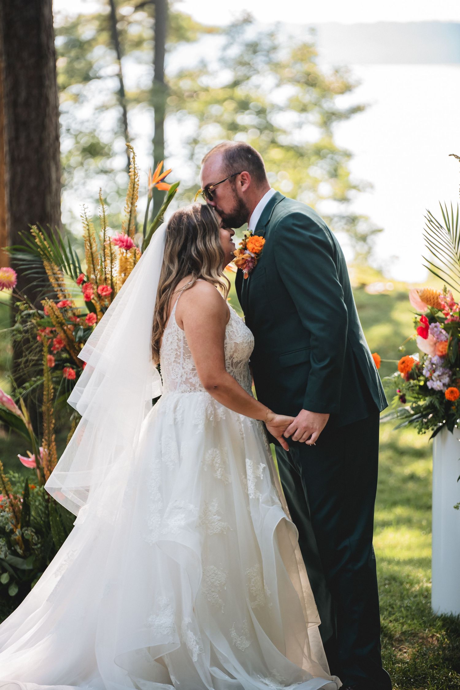 A romantic outdoor wedding ceremony moment with the couple sharing a kiss under floral arrangements at sunset.