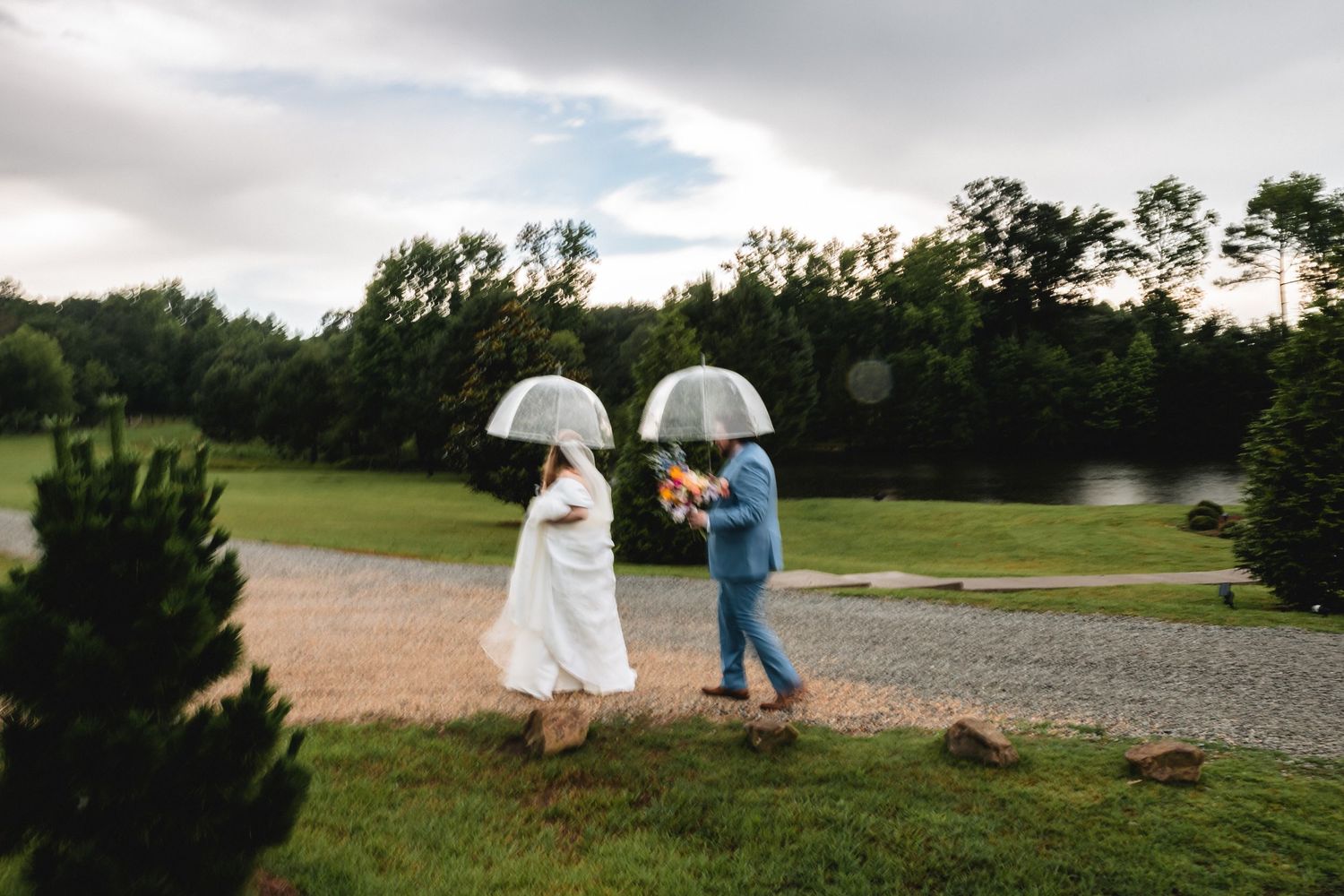 Bride and groom walking with clear umbrellas in rain at outdoor wedding venue with lush green trees.