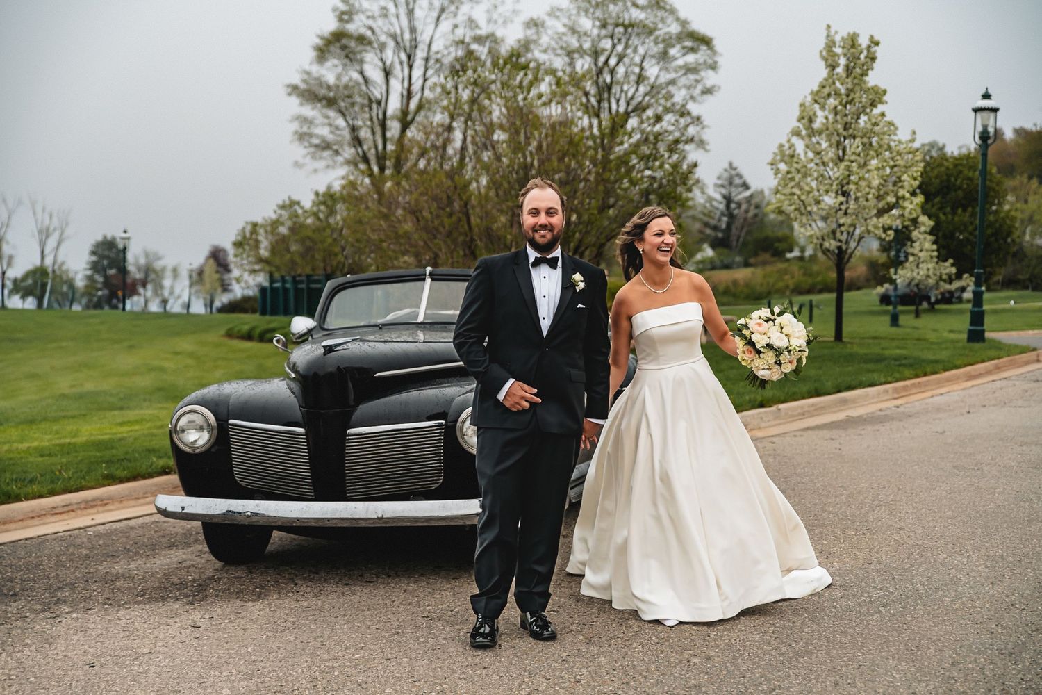 Bride and groom posing beside a vintage black classic car on a paved path at an elegant outdoor wedding venue.