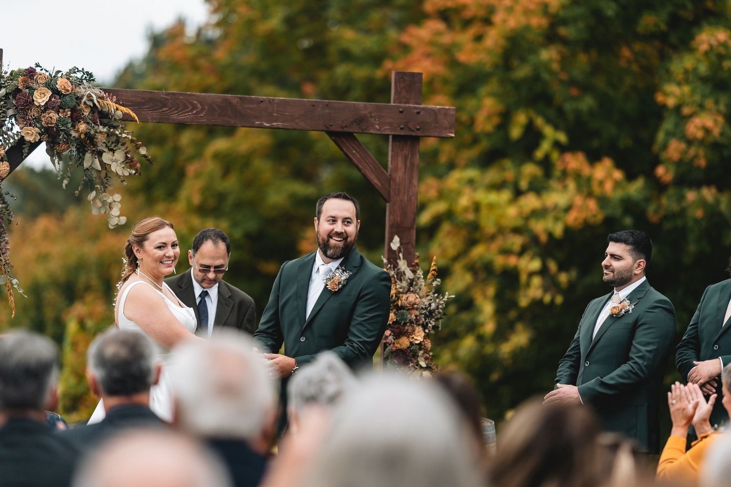 Outdoor autumn wedding ceremony with guests seated in white chairs surrounded by golden fall foliage.