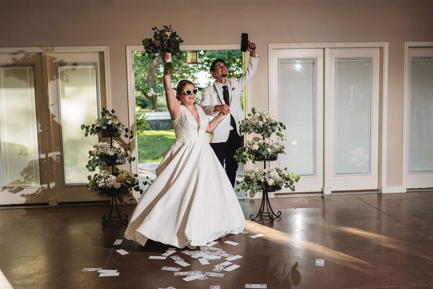 Bride and groom celebrate wedding entrance in sunglasses, confetti flying, surrounded by floral arrangements in elegant venue.