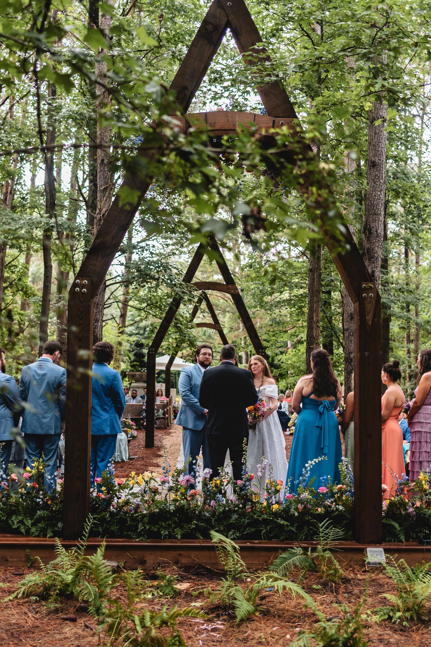 Outdoor forest wedding ceremony with couple at rustic wooden arch altar surrounded by wildflowers and guests in blue attire.