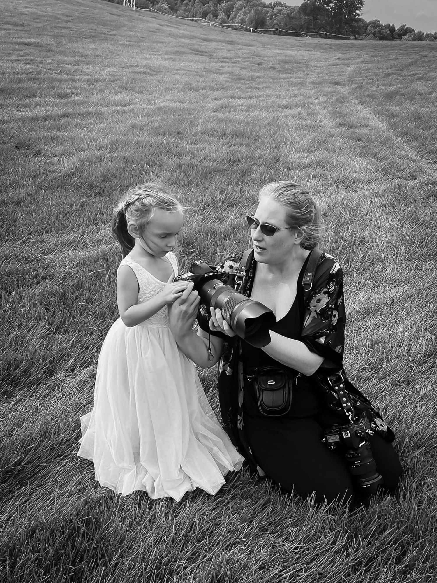 Black and white photo of a Northern Michigan wedding photographer showing a flower girl how to use a camera in a grassy field.