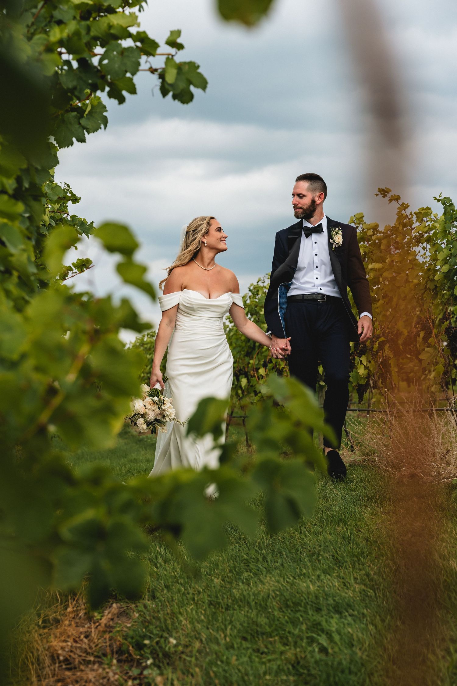 A bride and groom walk hand-in-hand through a lush vineyard, she in a white gown, he in a black tuxedo.