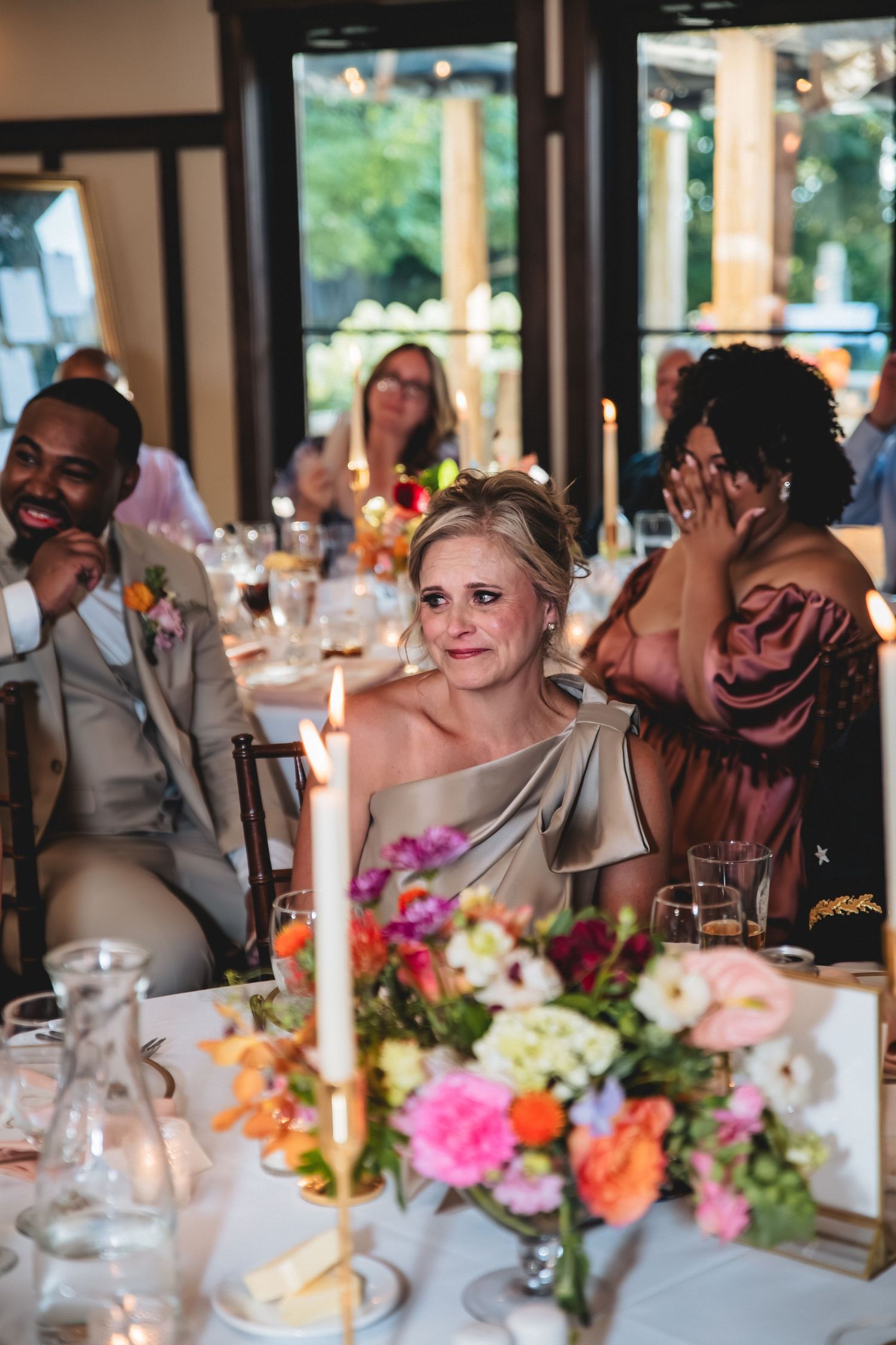Emotional wedding guest in gold dress seated at floral decorated reception table with candles and colorful centerpieces.