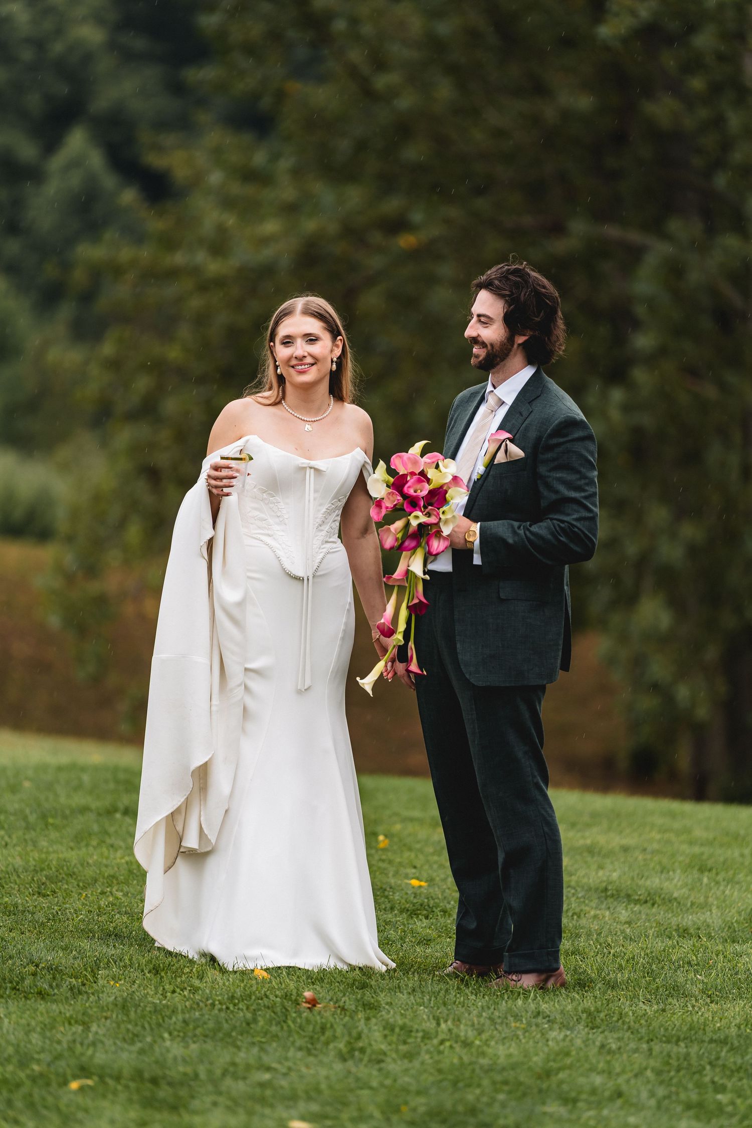Bride in white gown and groom in dark suit holding pink bouquet on green lawn with trees.