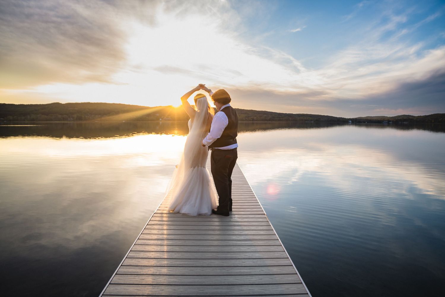 Two figures stand on a wooden dock overlooking a serene lake at sunset with dramatic clouds in the sky.