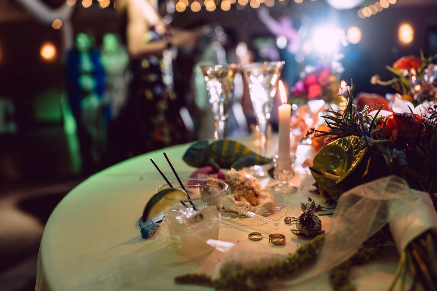 Elegant wedding reception table with floral arrangements, rings, and bokeh string lights in the background.