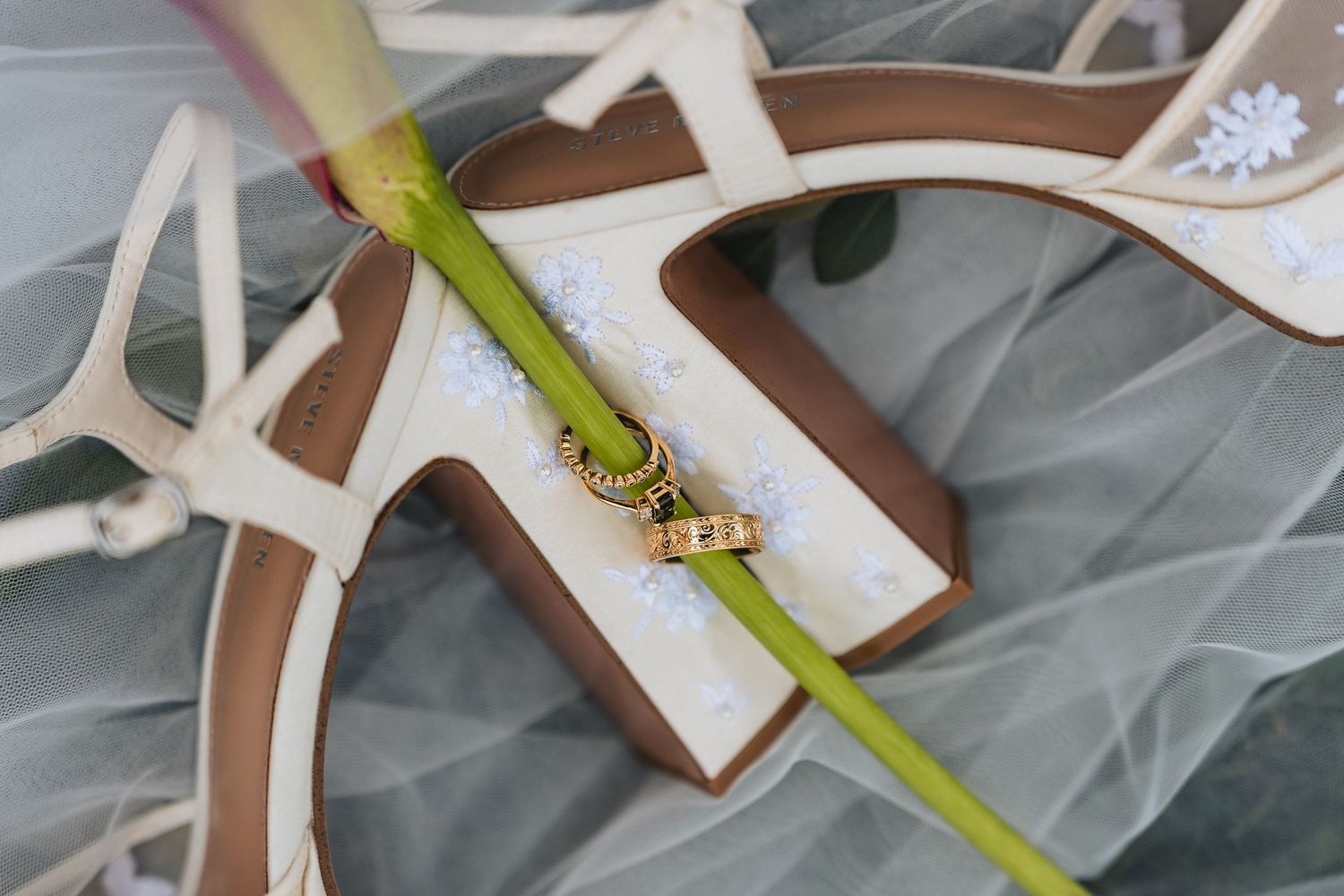 Wedding rings resting on white bridal heels with green stem and veil, elegant wedding detail photography captured by Treehouse Studios