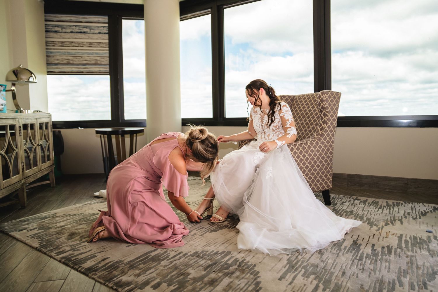 Bridesmaid in pink dress kneeling to help bride with shoes while bride sits in chair wearing white wedding gown.