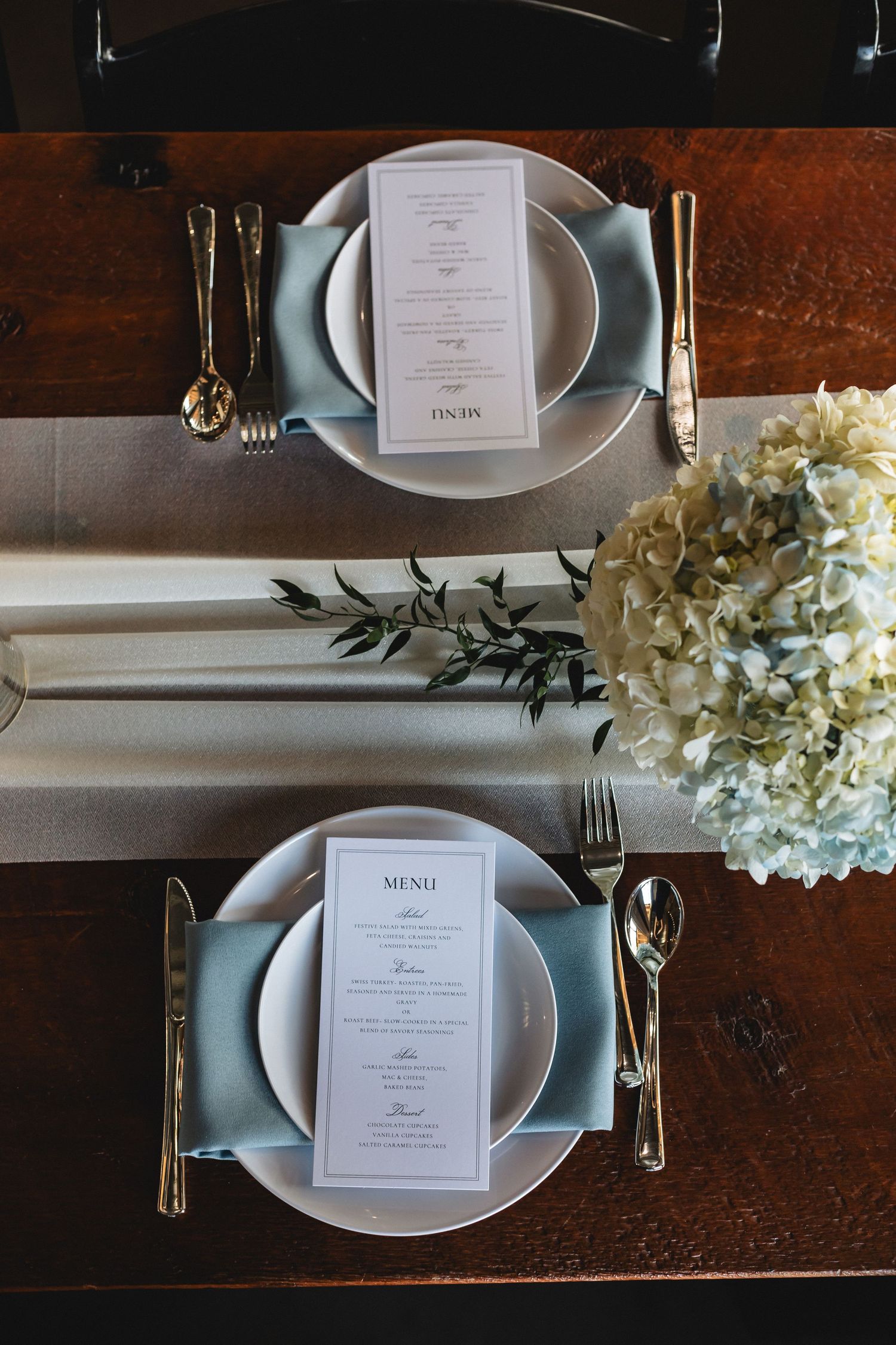 Elegant wedding table setting with gold cutlery, light blue napkins, white hydrangeas, and menu cards on a dark wood table.