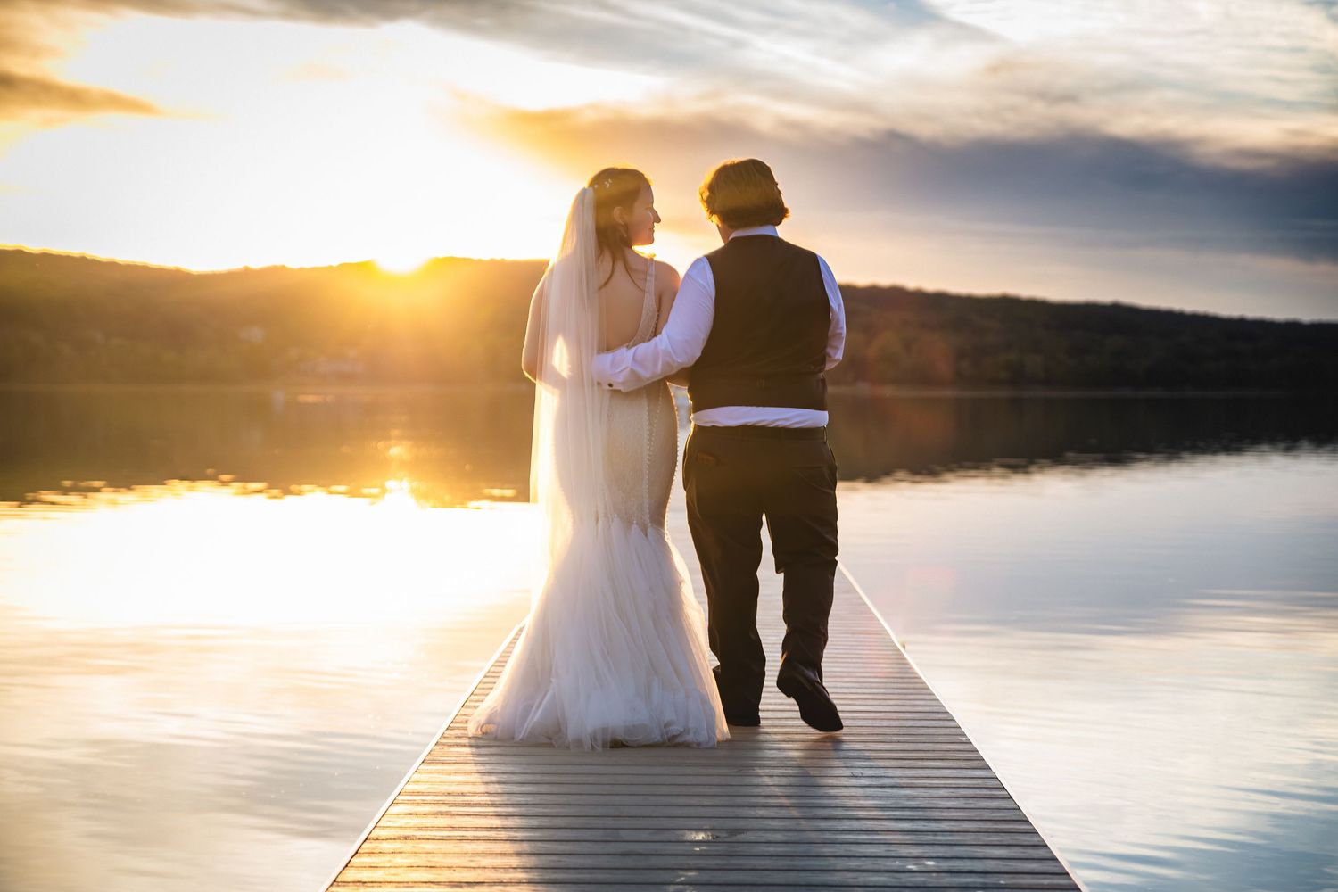 Bride and groom walking on a dock at sunset, with golden light reflecting on calm lake waters.