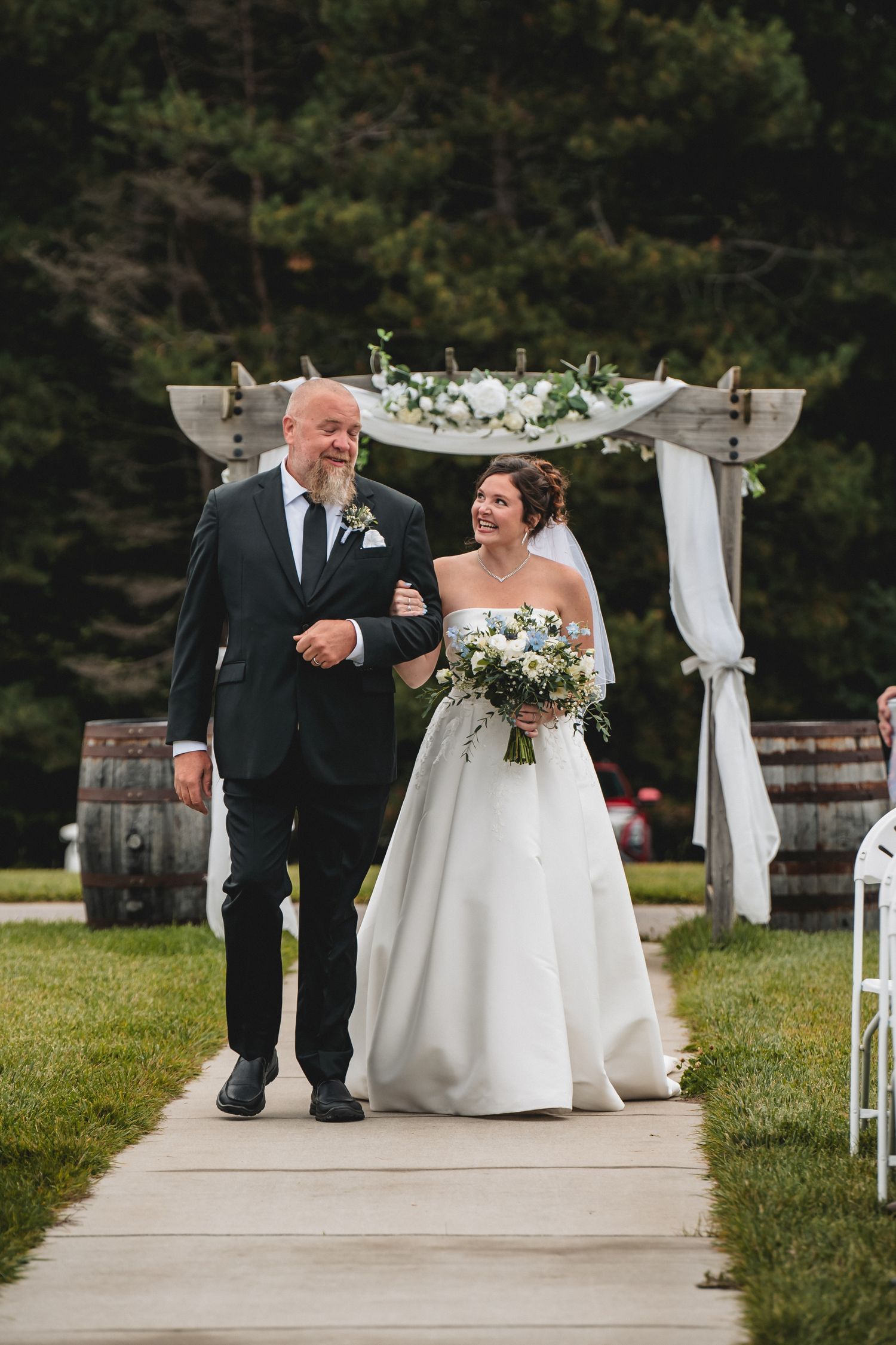 Bride in white gown walks down outdoor aisle with older man in dark suit, holding white floral bouquet under decorated arch.