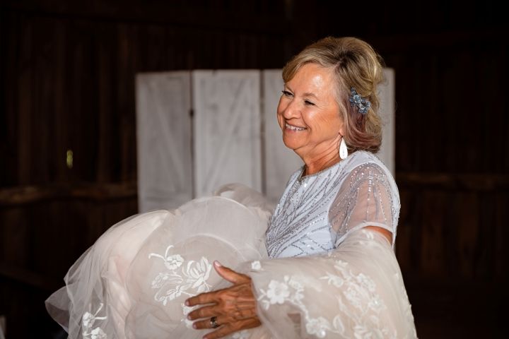 Smiling woman in elegant lace gown holding a wedding dress in a rustic barn setting.