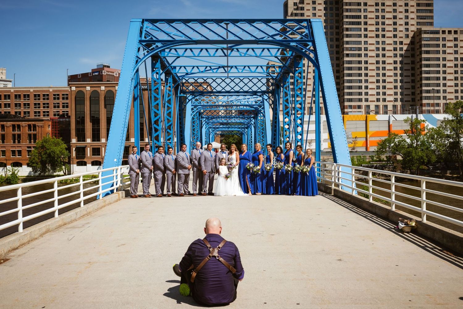 Photographer sits on blue bridge capturing wedding party in blue attire with city skyline backdrop.