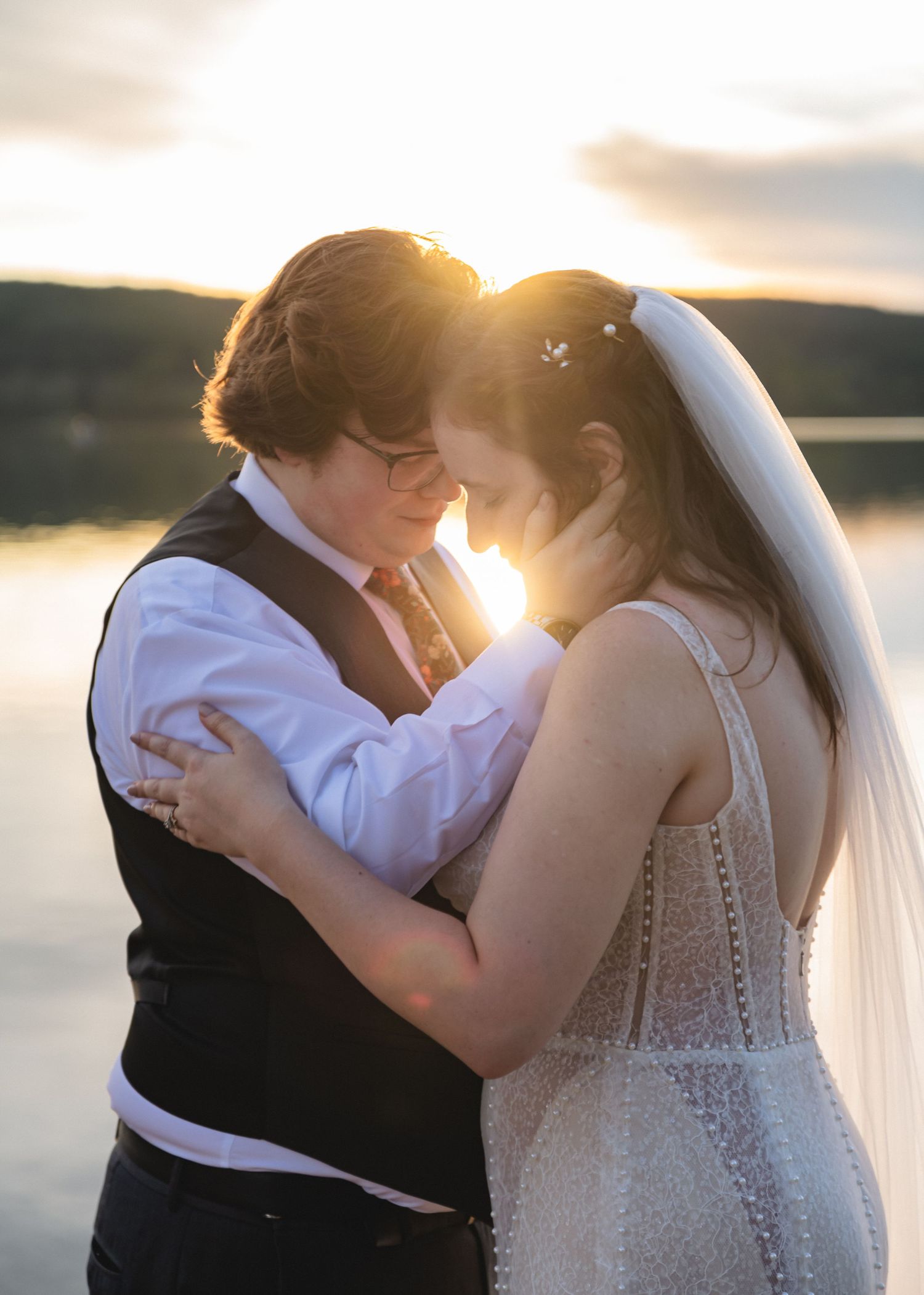 Couple in wedding attire sharing a romantic sunset moment by a lake, bride in veil and groom in vest.