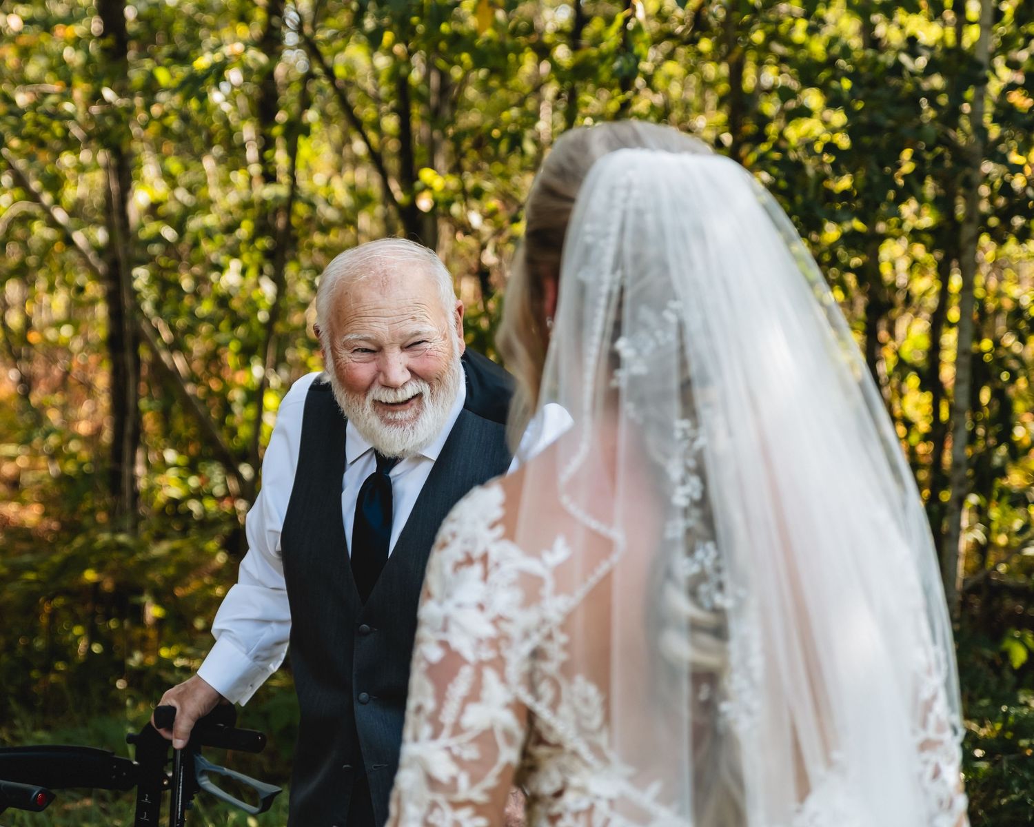 Elderly man with white beard smiles joyfully at bride in lace wedding dress and veil outdoors.