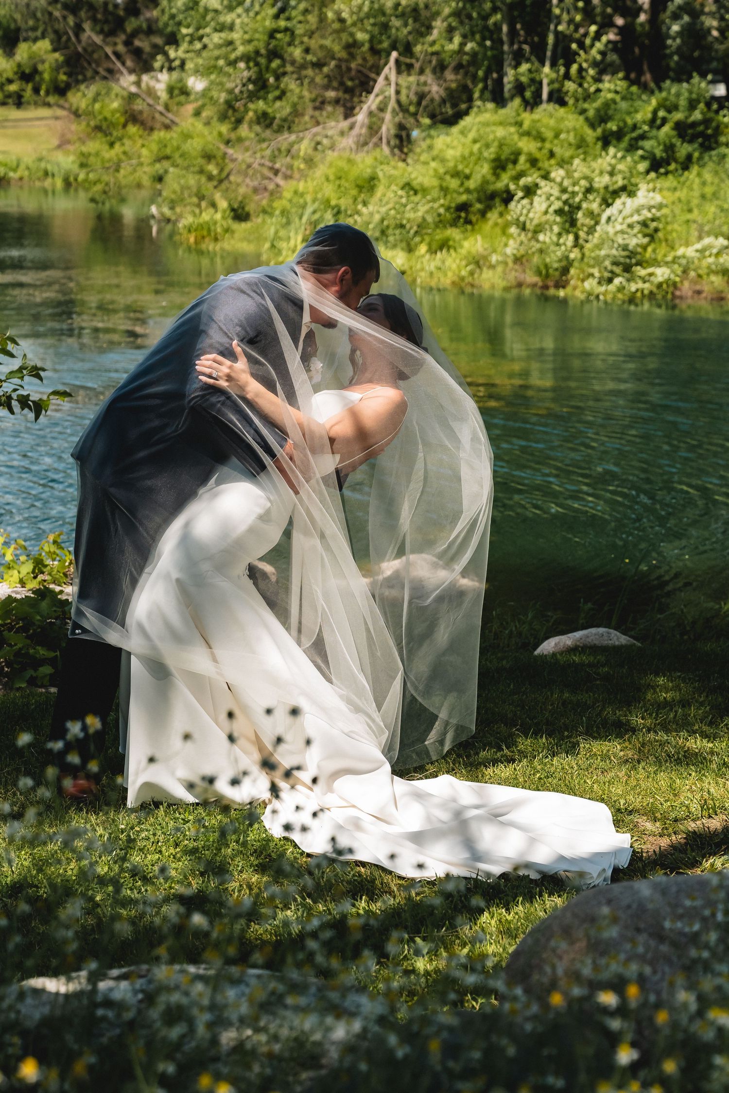 Bride and groom sharing a romantic kiss under a flowing veil by a serene green lake on their wedding day.