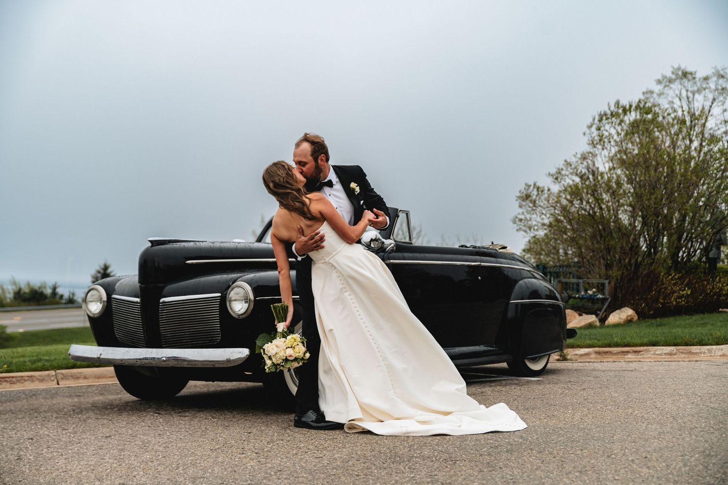 A couple shares a romantic dip pose in front of a classic black vintage car on their Northern Michigan wedding day.