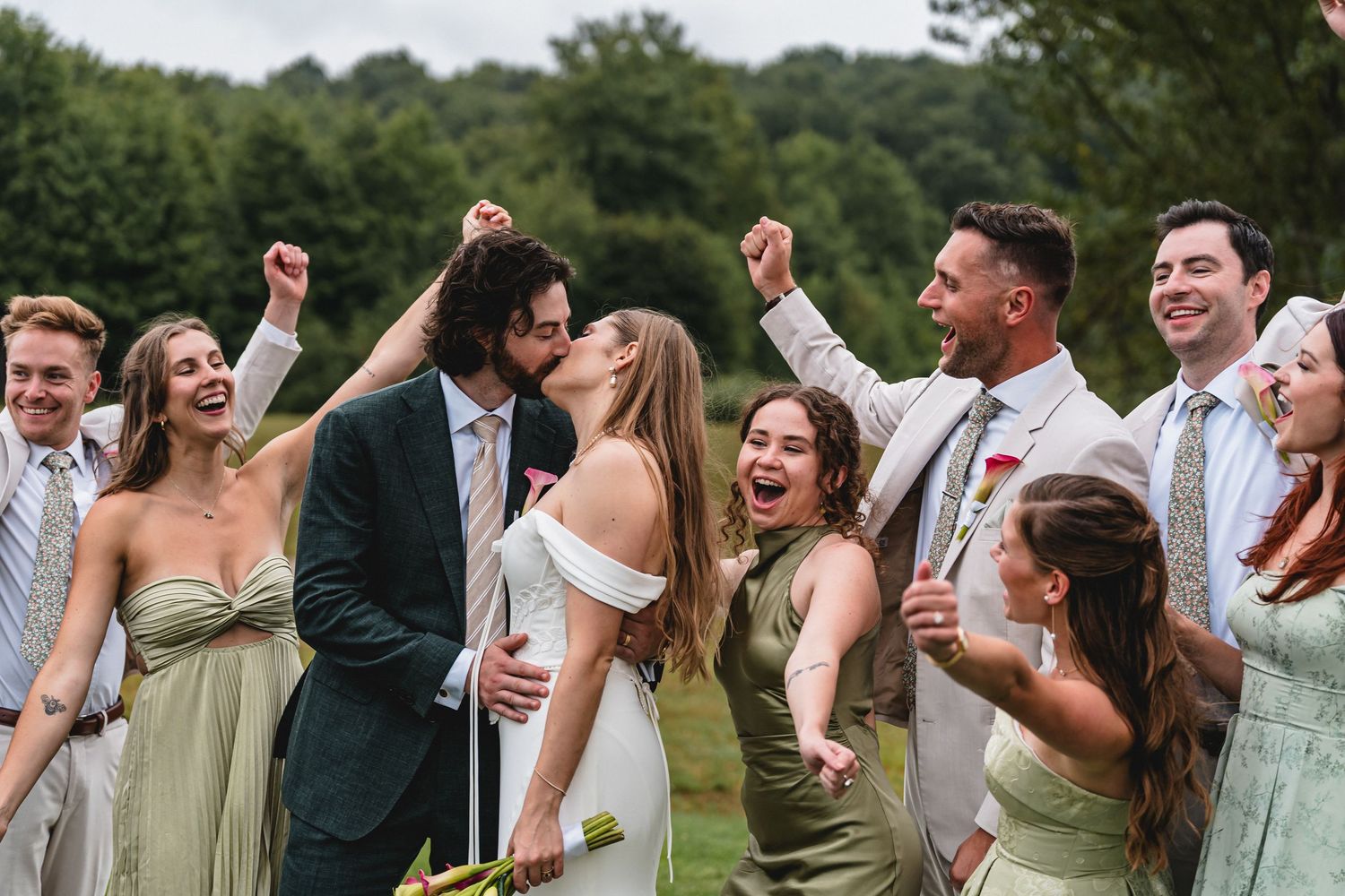 Joyful wedding party celebrates as bride and groom kiss outdoors, surrounded by cheering bridesmaids and groomsmen.