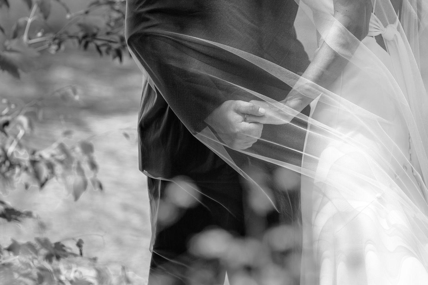 Black and white wedding photo of bride and groom holding hands, soft veil draped over couple in garden setting.