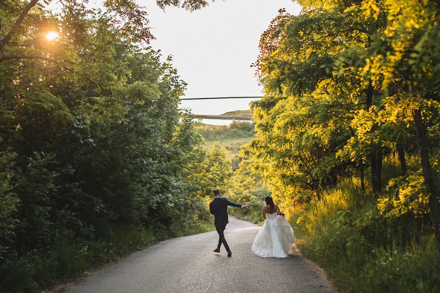 Bride and groom walking hand in hand down a sunlit tree-lined road during golden hour wedding photography.