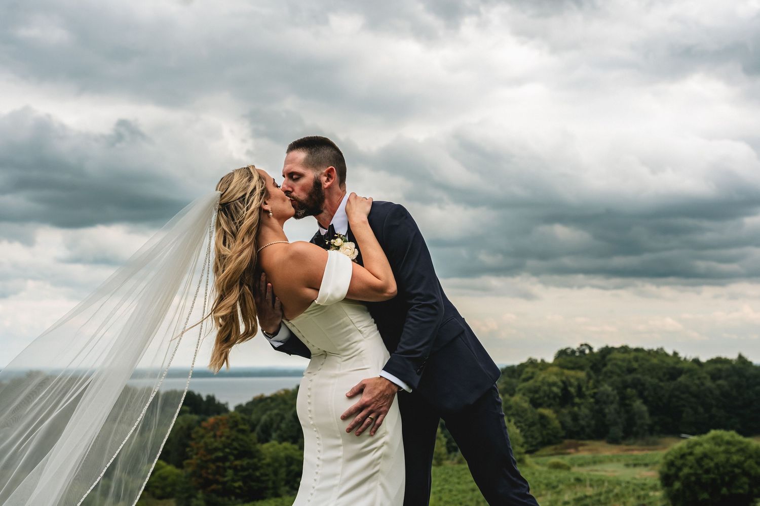 Bride and groom share romantic kiss outdoors with dramatic cloudy sky and vineyard landscape at Chateau Chantal Mission Peninsula