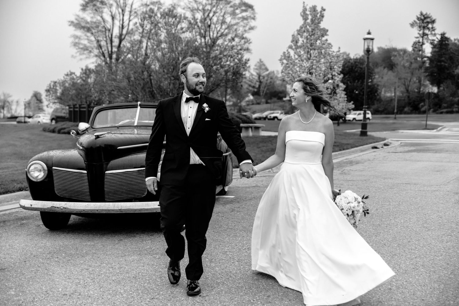 A joyful couple walks together on their Northern Michigan wedding day with a vintage black car in the background.