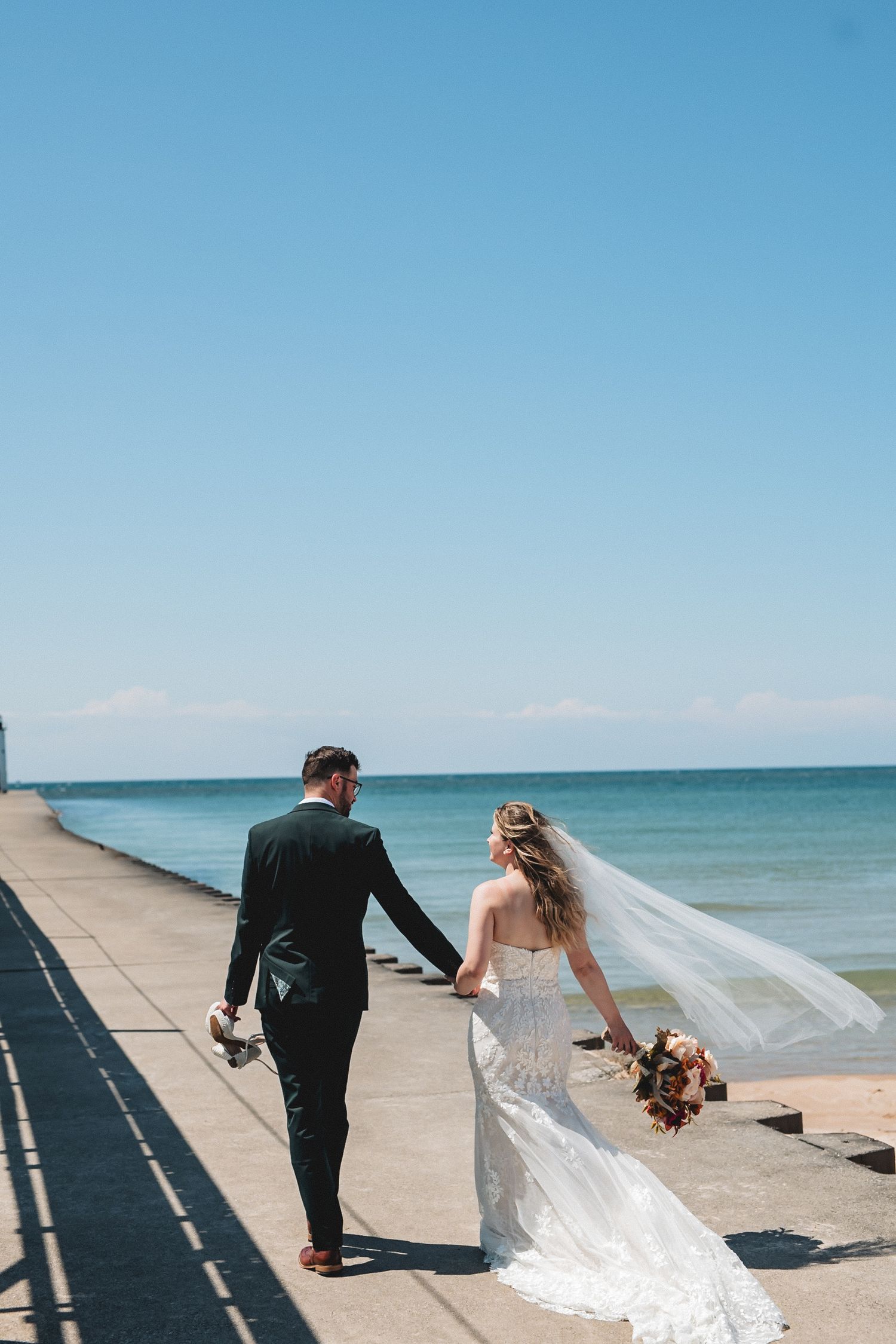 A couple in wedding attire walks along a waterfront promenade with the bride's veil flowing in the breeze.