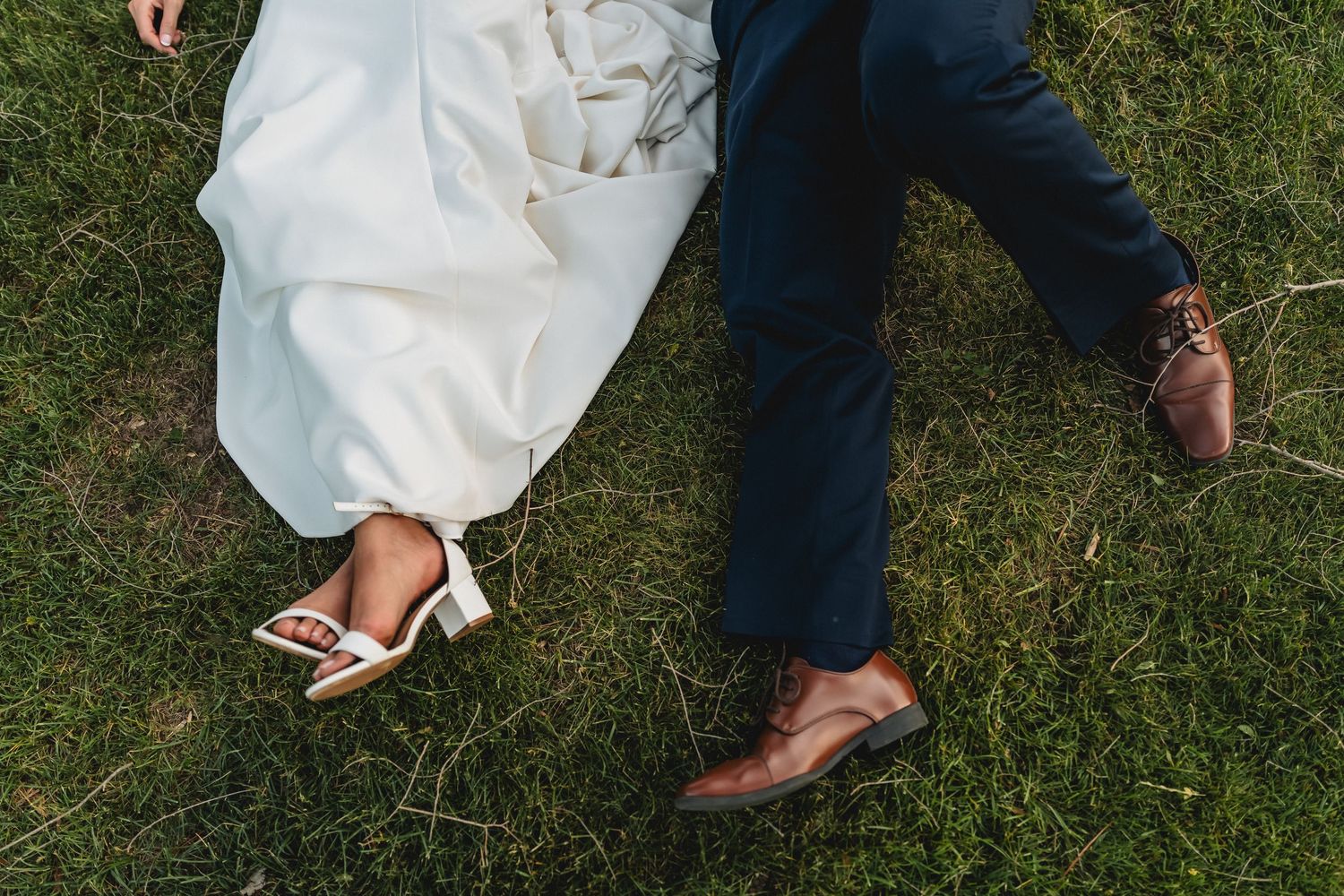 Bride and groom lying on green grass, showing white wedding heels and brown leather dress shoes with navy suit pants.
