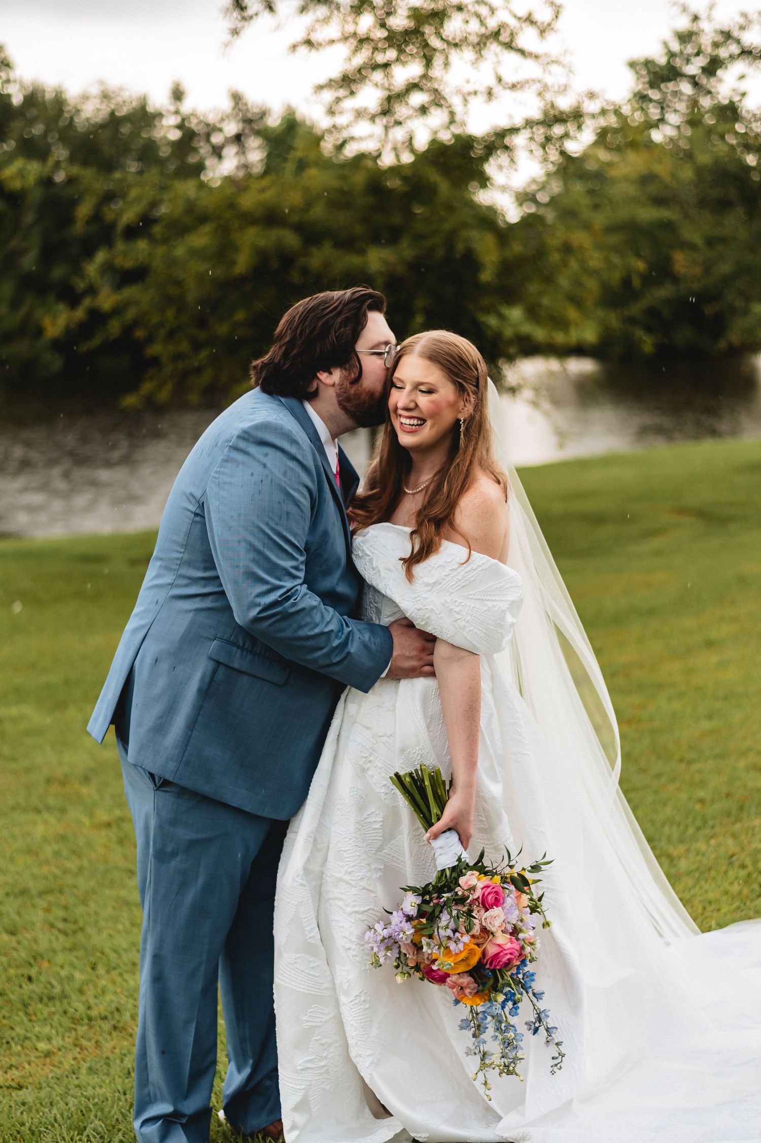 A romantic wedding couple embraces on a grassy lawn during a sunset photoshoot with a colorful bouquet.