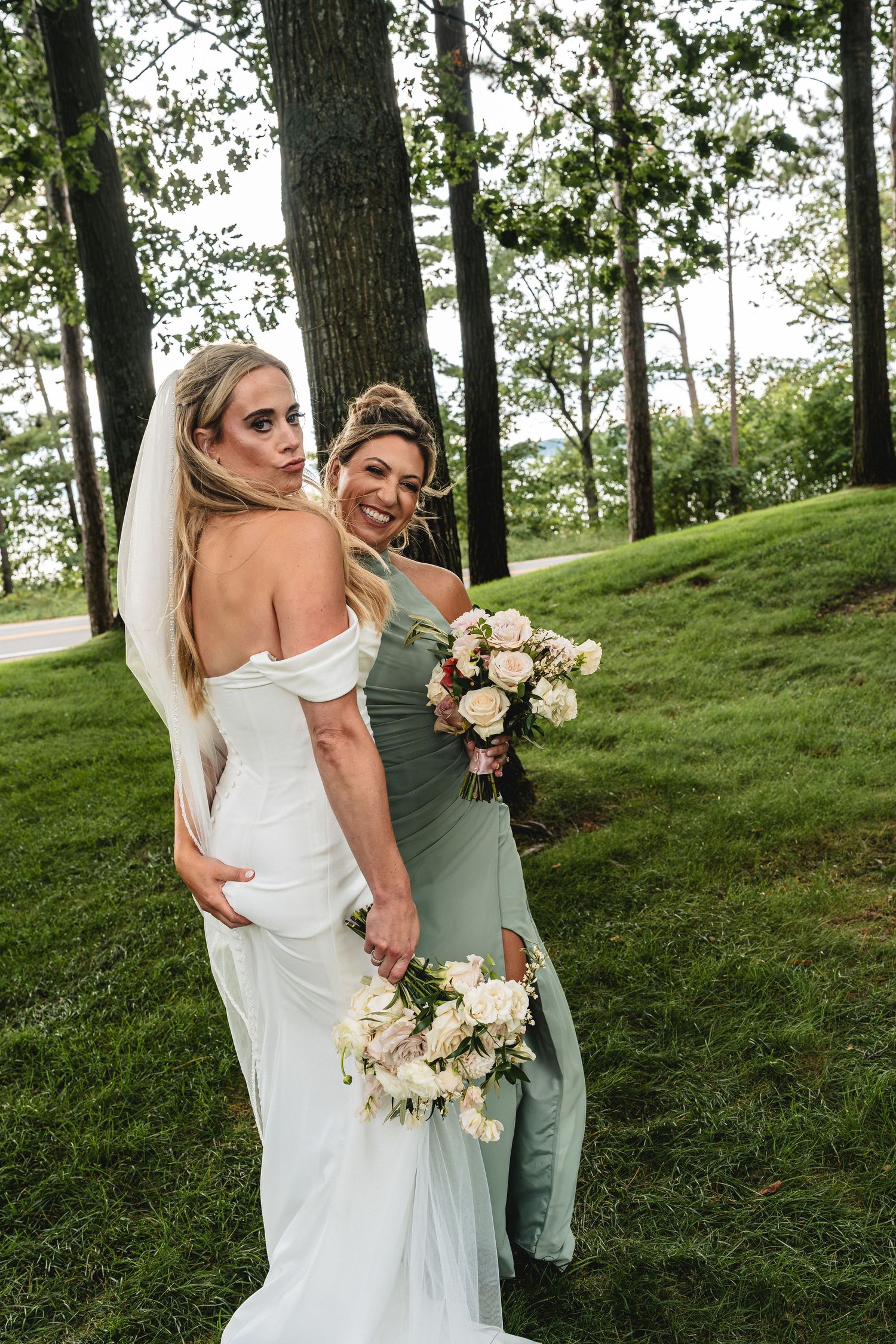Bride poses with smiling bridesmaid in sage green dress, both holding bouquets at a nature inspired northern Michigan destination wedding