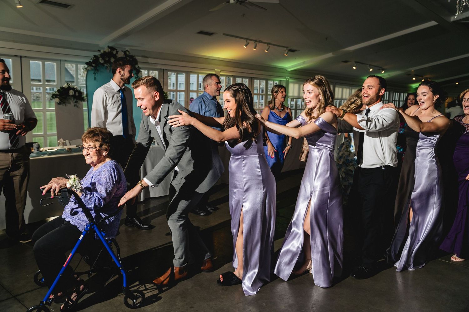 Wedding guests in purple bridesmaid dresses dance joyfully at a reception, including a woman using a walker.