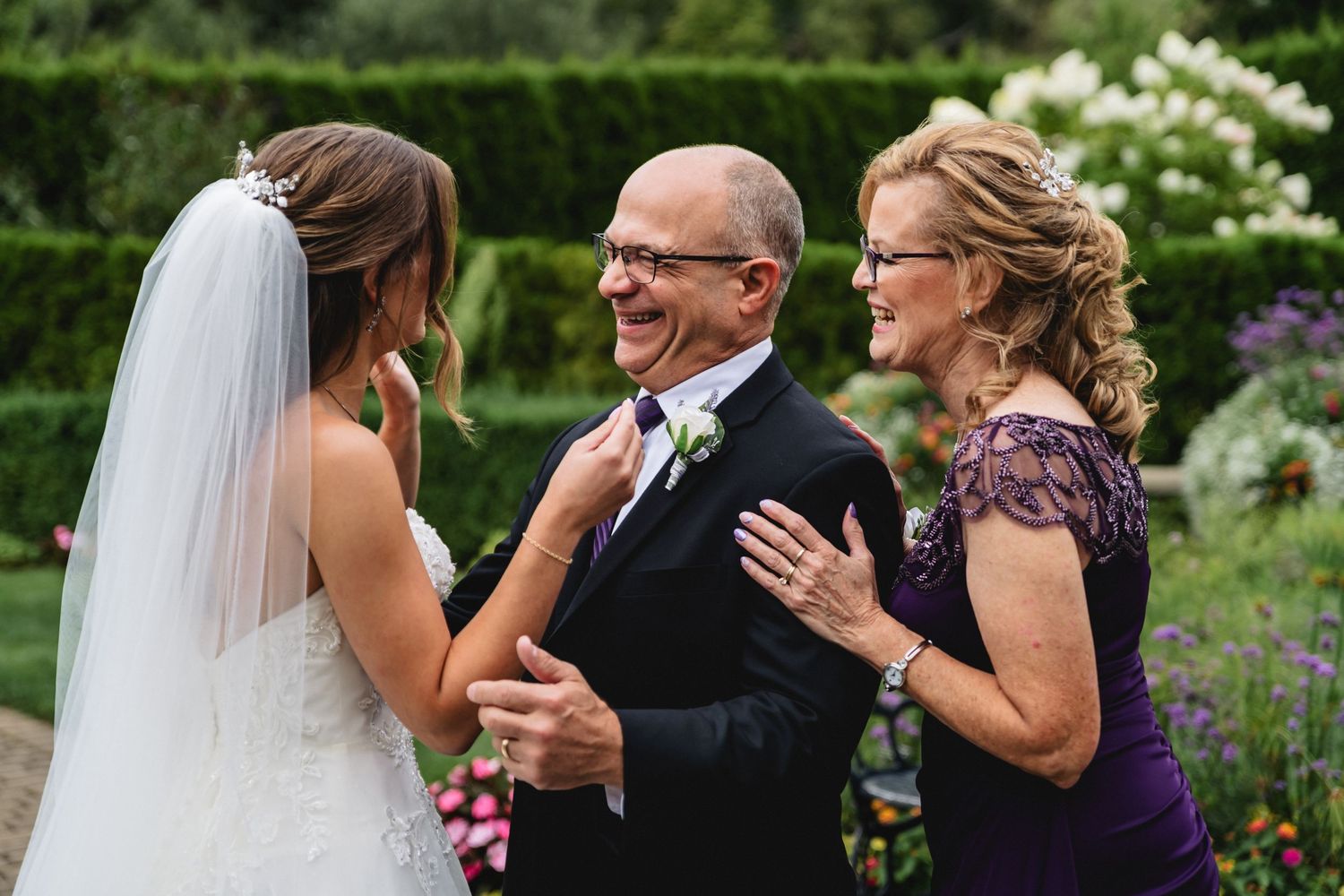 Bride shares a joyful moment with smiling parents in formal attire at an outdoor garden wedding ceremony.