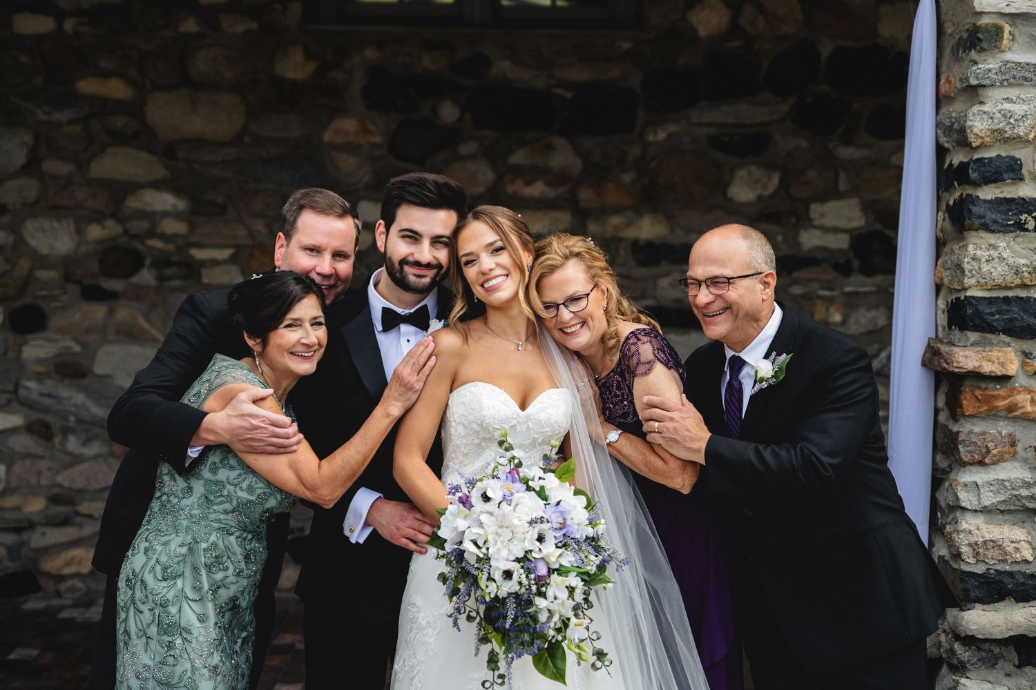 Wedding party laughs and embraces while posing against rustic stone wall during outdoor celebration.