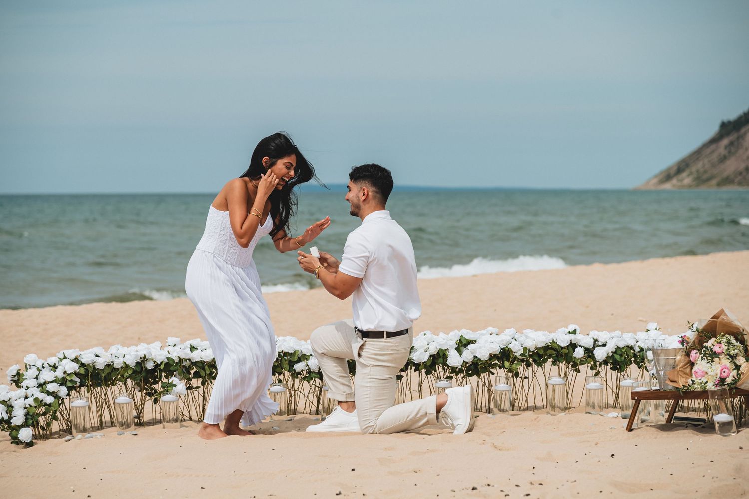 Man proposes to woman on a sandy beach with white flowers, ocean backdrop, both dressed in white.