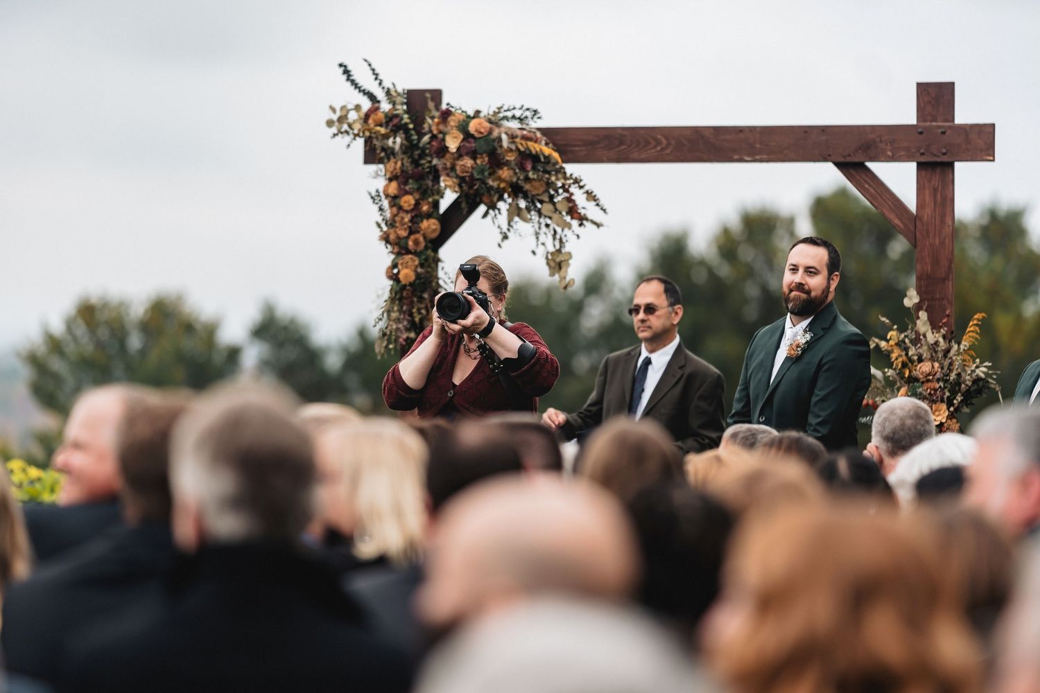 Outdoor wedding ceremony with guests seated, officiant and groom at rustic wooden arch adorned with autumn floral arrangements.