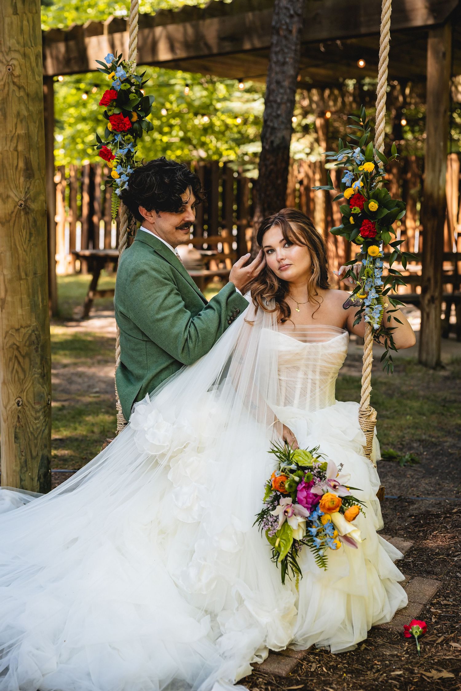 Bride in white ruffled gown sits on floral swing as groom in green suit tenderly touches her hair outdoors.