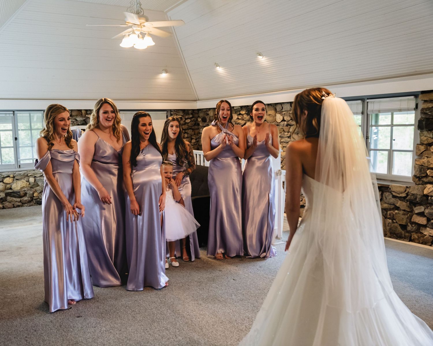 Bridesmaids in lavender satin gowns react with joy as bride in white wedding dress and veil reveals her look indoors.