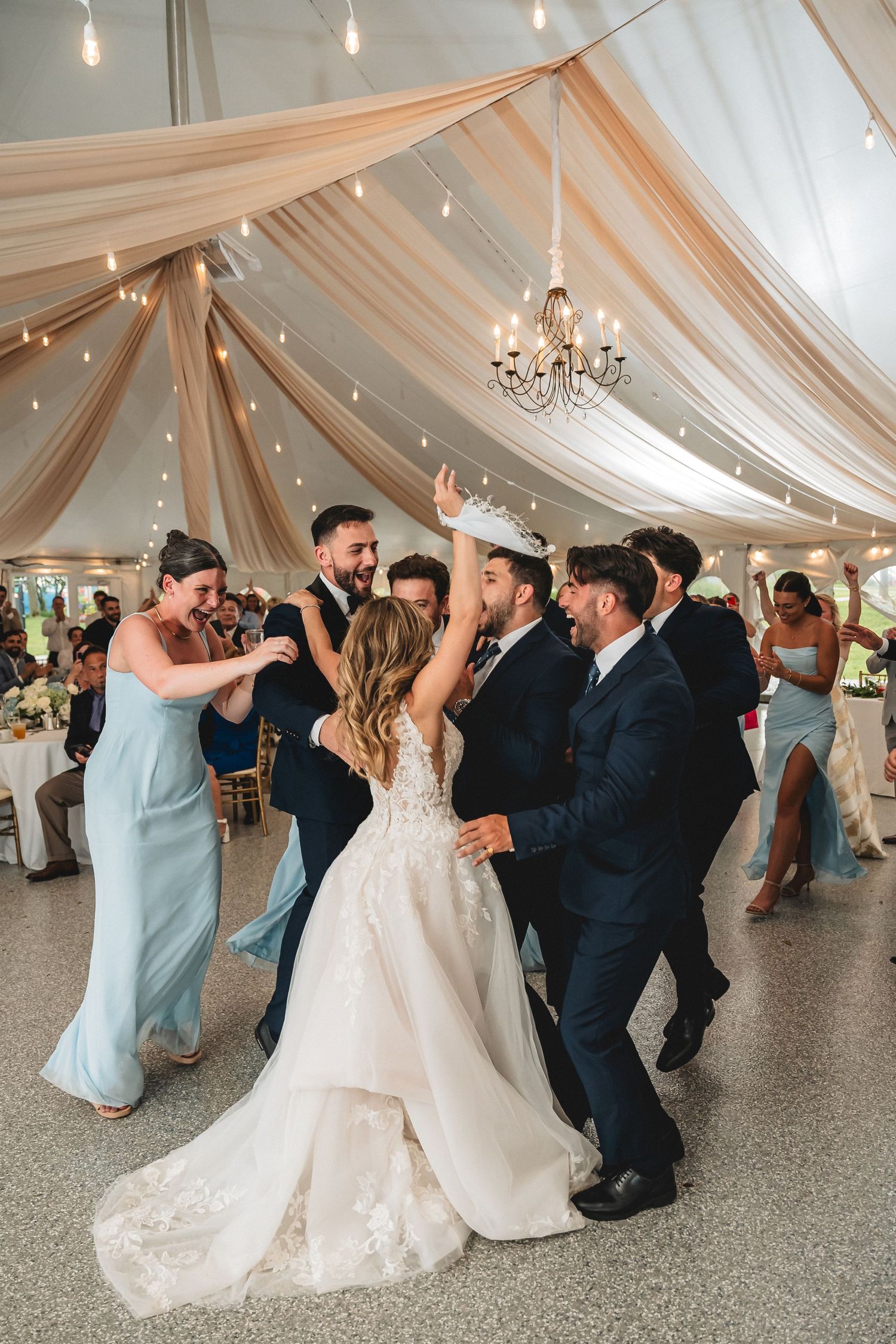 Bride and groom dancing joyfully with wedding party under elegant draped tent with string lights.