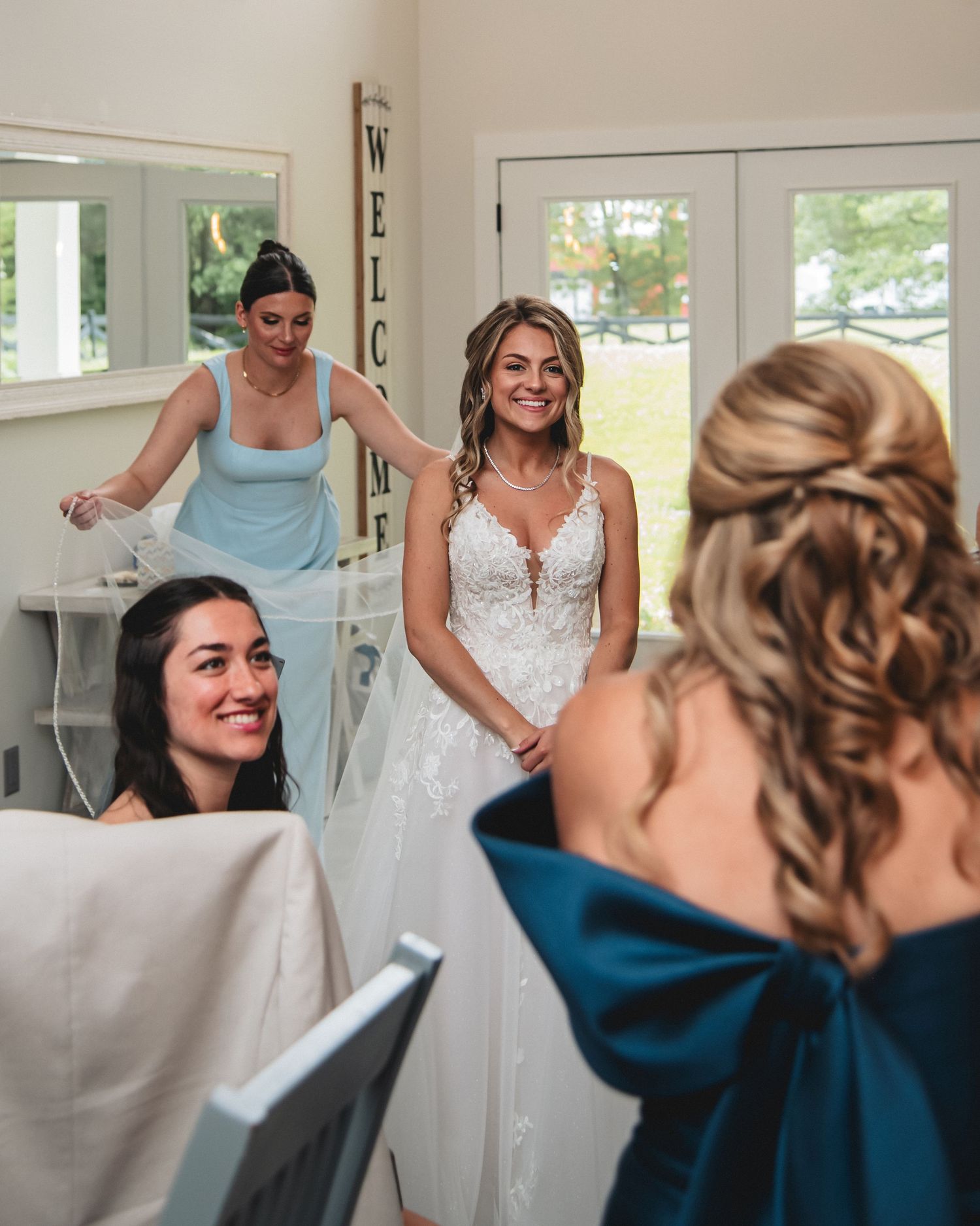 Bride in white lace gown smiles at bridesmaids in blue dresses during wedding preparations indoors.
