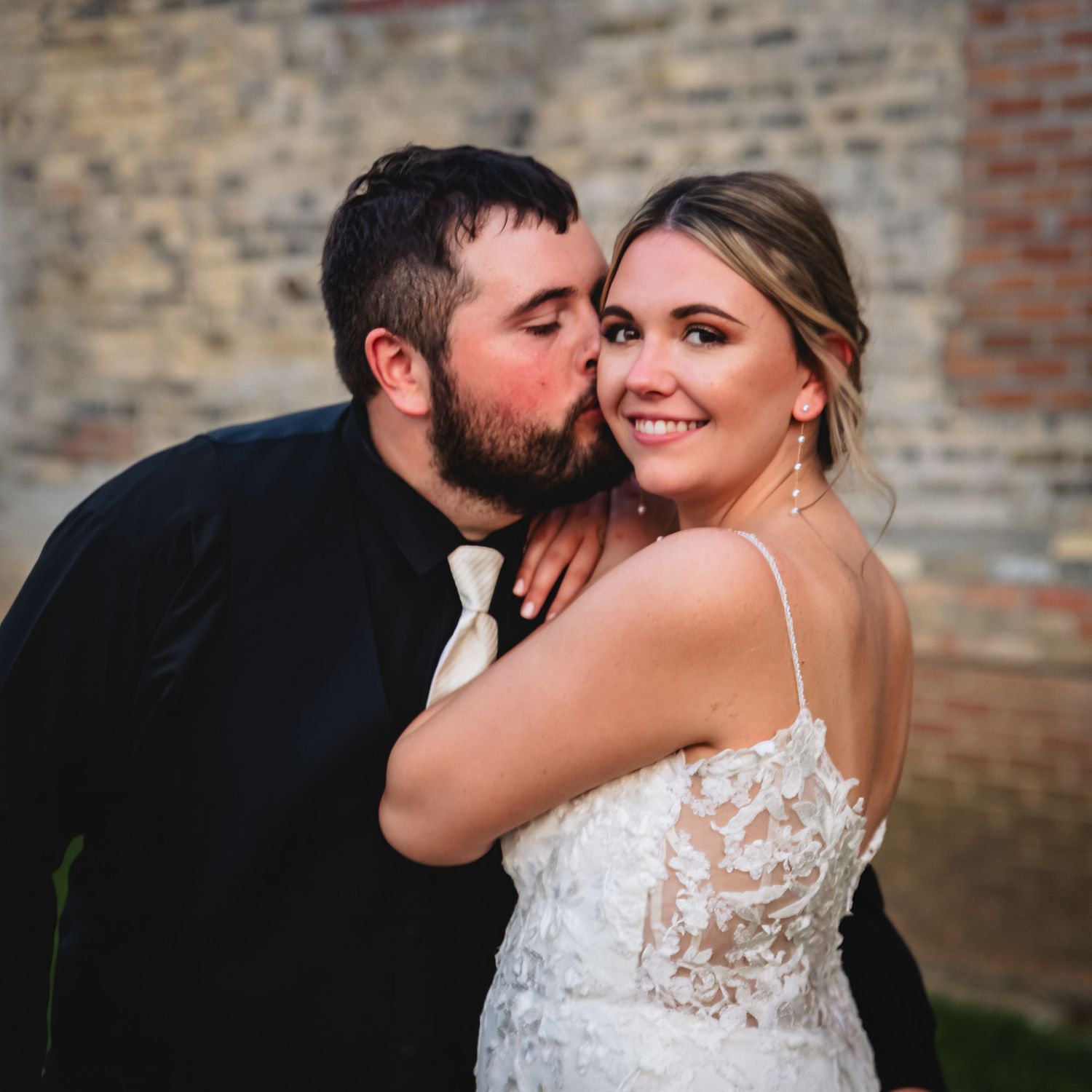 Bride and groom share a romantic kiss on cheek pose in front of rustic brick wall at outdoor wedding.