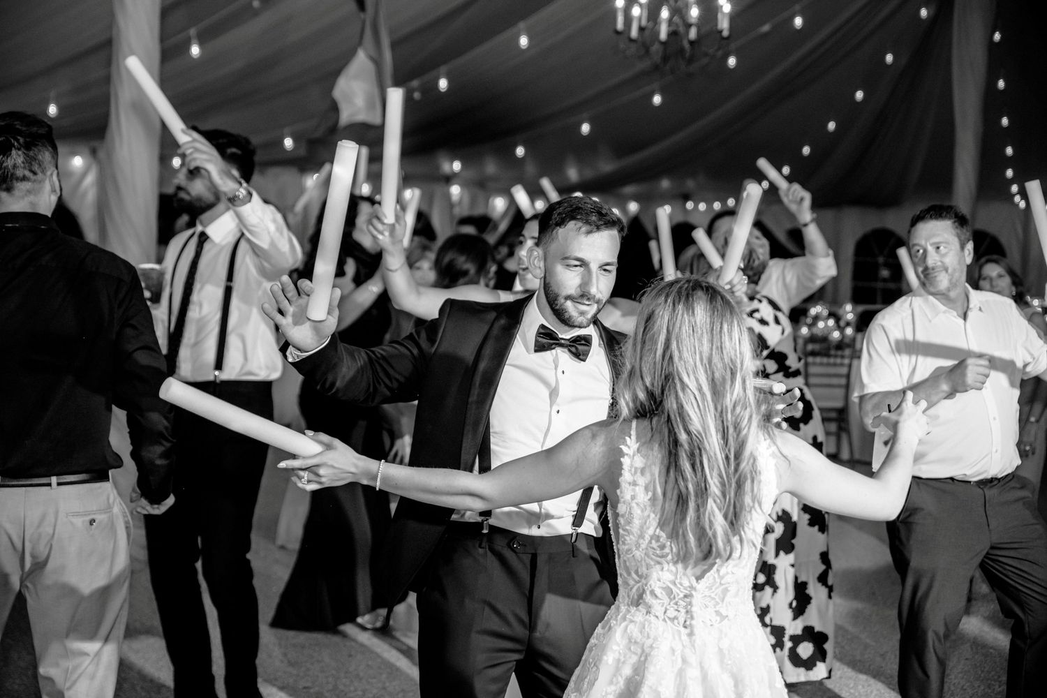 Bride and groom dancing with glowing sticks at wedding reception under tent with string lights.