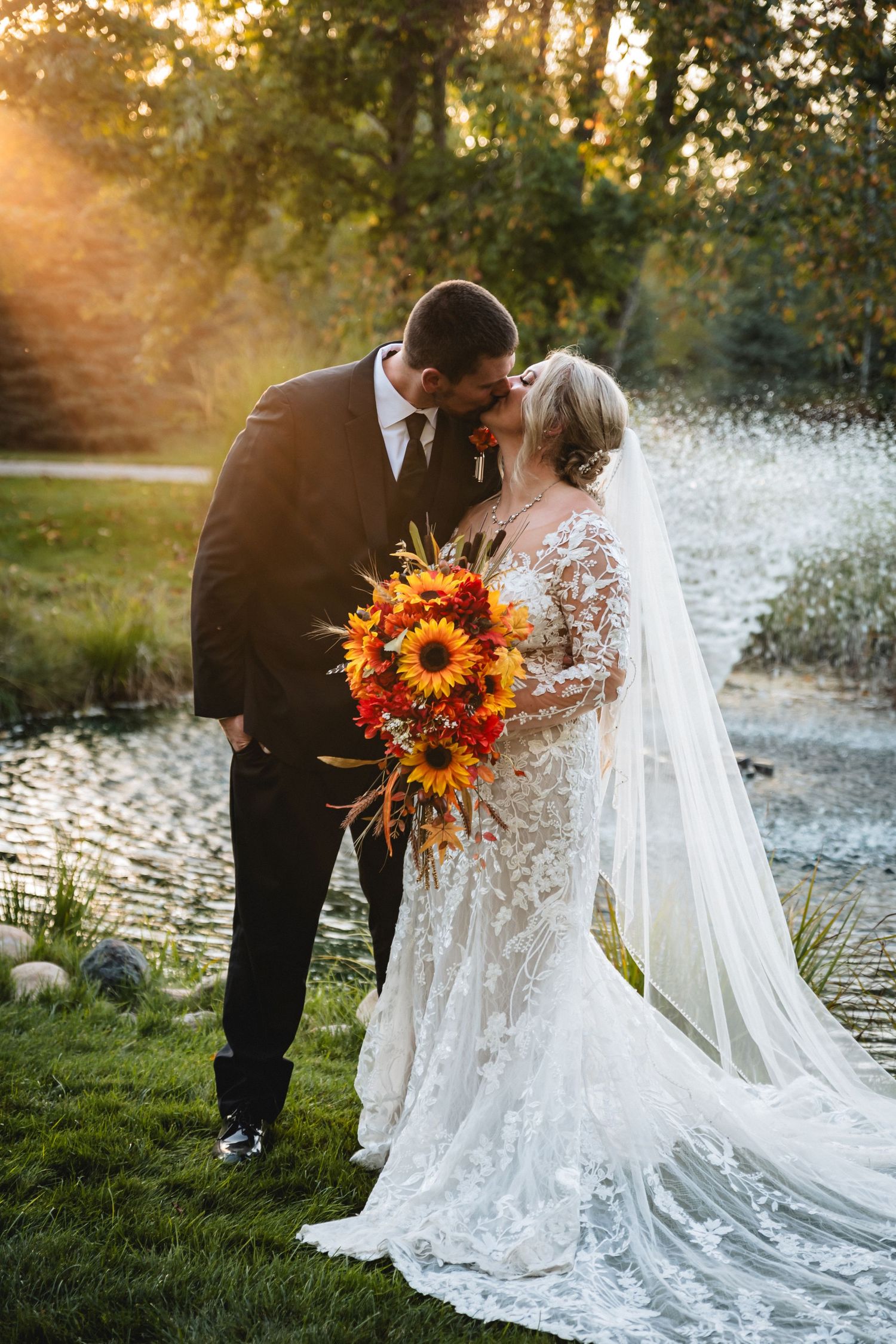 Bride and groom share a kiss at golden hour by a pond, bride holding vibrant sunflower bouquet in lace gown.