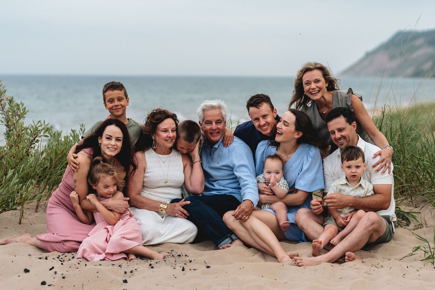 Multi-generational family smiling together on a sandy beach, with ocean waves and green hills in the background.
