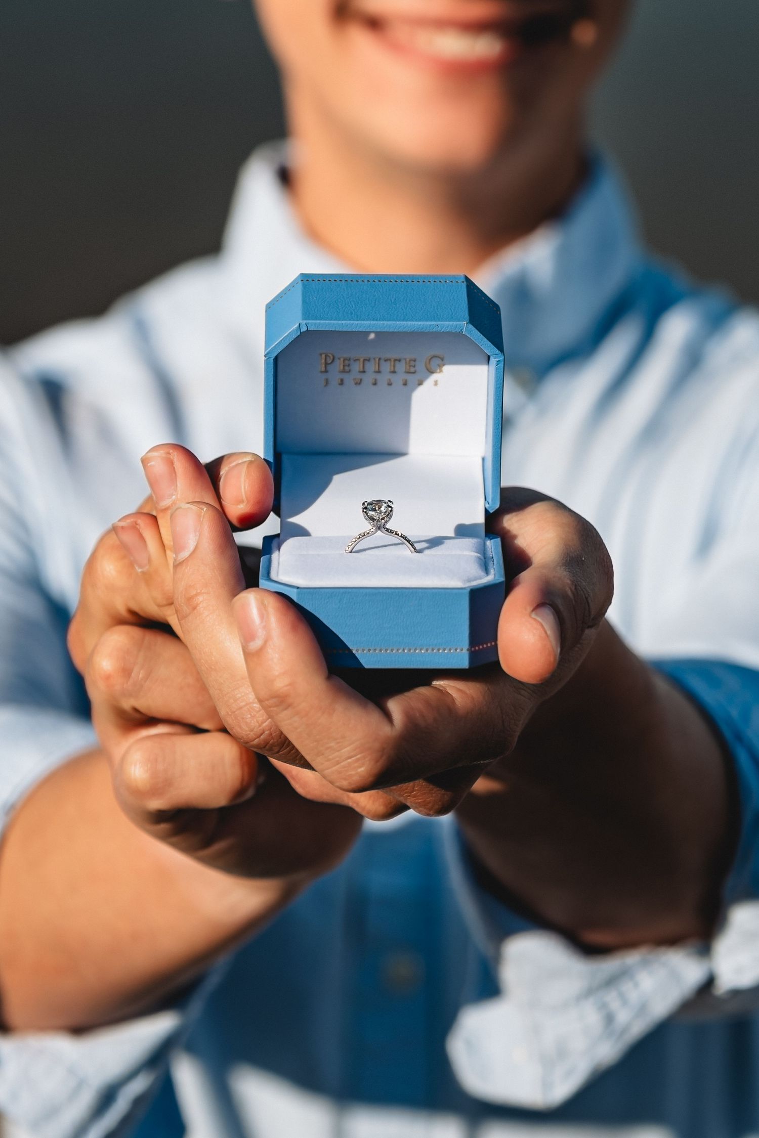 Smiling man in denim shirt presenting an open blue Petites jewelry box with a diamond engagement ring.
