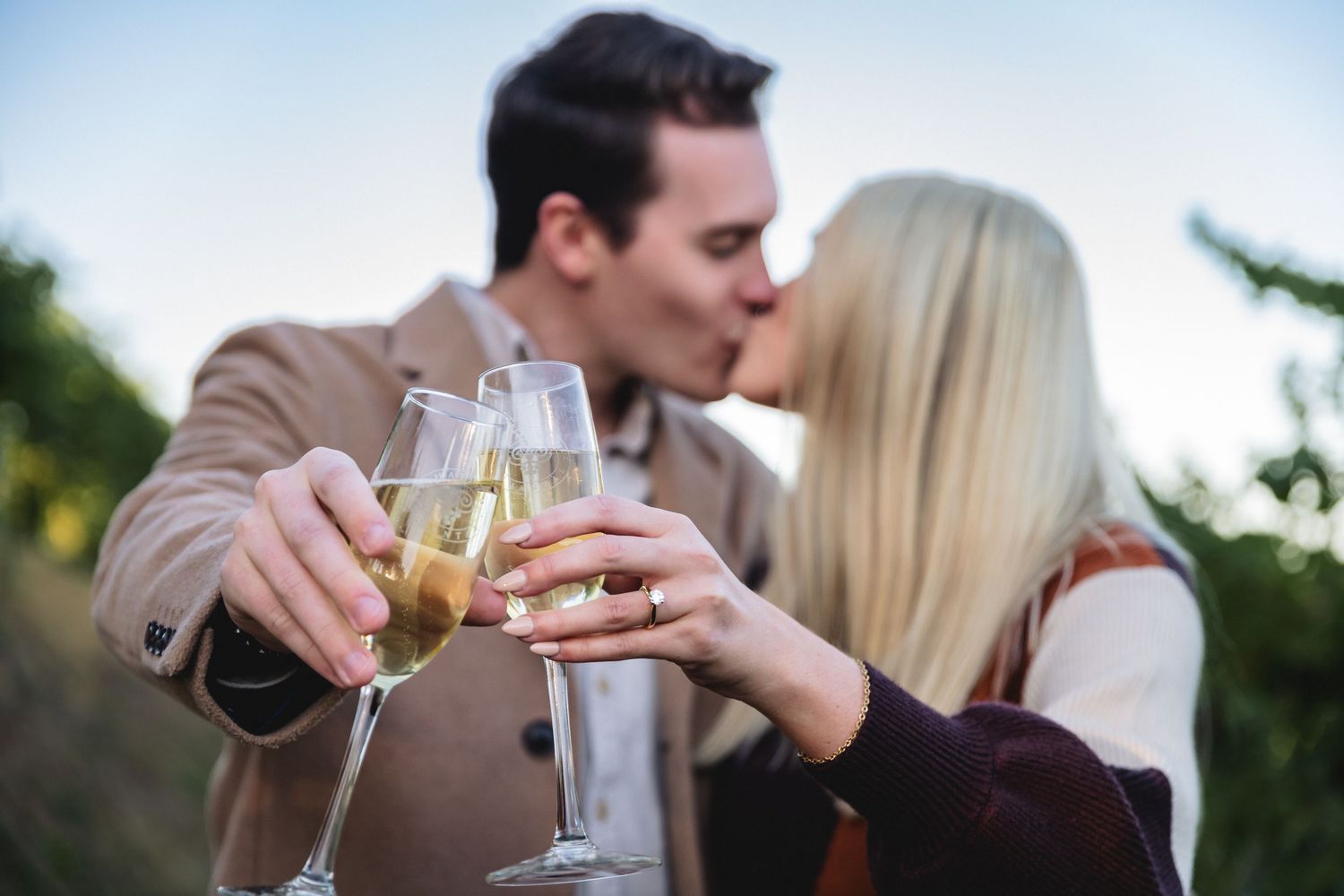 Couple kissing while clinking champagne glasses outdoors, celebrating a romantic moment together.